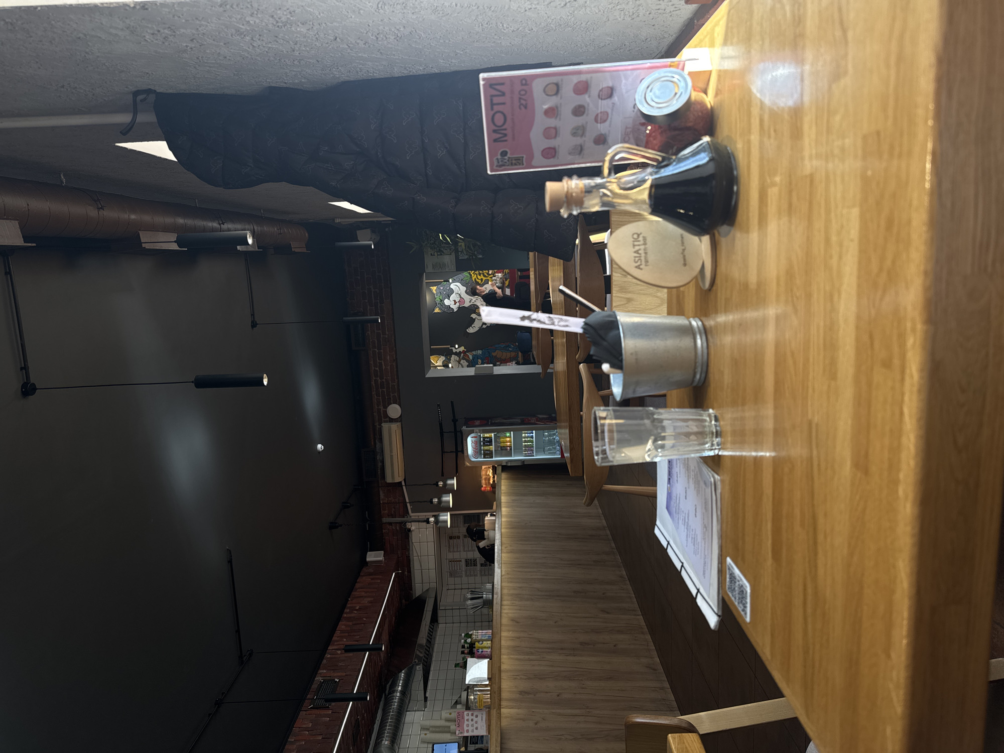 This media file presents an interior view of a modern, casual restaurant or ramen bar in Saint Petersburg, Russia. The perspective is from a diner's seat, looking across a light-colored wooden table towards the back of the establishment.
On the wooden table in the foreground, dining accessories are arranged: a small glass carafe holding a dark liquid, likely soy sauce, and a round glass jar with a metallic lid containing a condiment. A white coaster with "ASIATIQ ramen-bar" printed on it is visible, along with a silver metal bucket holding chopsticks and two empty clear glasses. A menu or flyer displaying a QR code also rests on the table.
Beyond the table, the restaurant's interior features a dark, industrial aesthetic with exposed brown-toned pipes running along the ceiling and walls. Track lighting fixtures illuminate the space. A dark puffer jacket hangs from a hook on a wall. A menu board prominently displays "MOTI" and a price of "270 P" (Rubles), showing various mochi options. Further inside, a service or kitchen counter with white tiled walls is visible. A large, stylized cat mural or decor piece adds a distinct artistic touch to a wall. A commercial refrigerator stocked with drinks, including Coca-Cola products, is positioned in the background. A staff member is partially visible behind the counter.
The scene is brightly lit, suggesting it is daytime. The combination of industrial design elements, specific menu items like mochi and ramen, and the visible Russian currency and text strongly indicate its location in Russia, aligning with Saint Petersburg. The overall atmosphere is functional, clean, and contemporary.