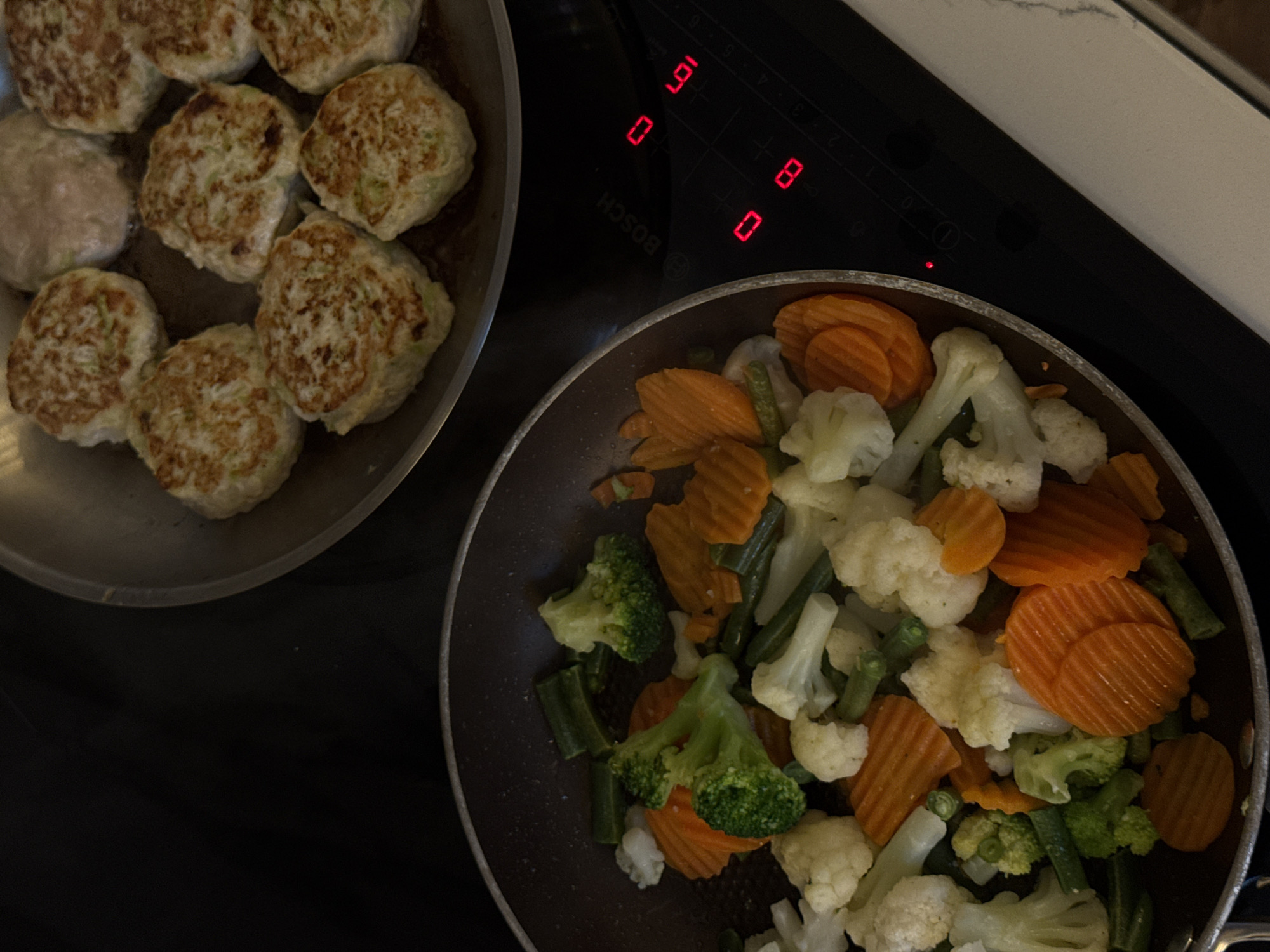 The image depicts a cooking scene on a black Bosch induction cooktop. Two pans are present: one on the left contains cooked patties, with some appearing golden brown and others lighter in color. The pan on the right contains a mix of cooked vegetables, including sliced carrots, broccoli florets, cauliflower florets, and green beans. The cooktop display shows a '9' on one burner and '0' on another. The scene is set on a countertop with a white, patterned surface visible in the upper-right corner. Moscow, Russia is not evident from the image content.