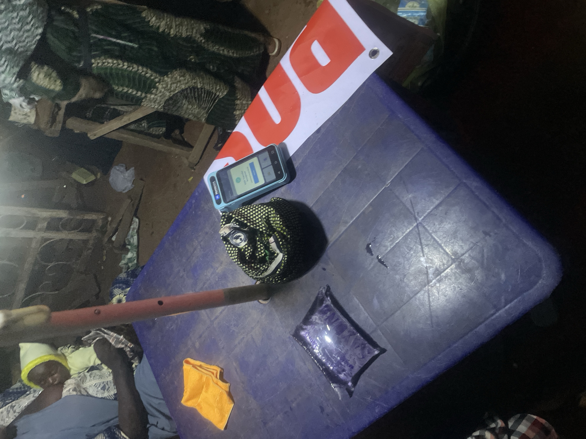 The image shows a close-up view of a blue plastic table with various objects on it. In the foreground, a person wearing a blue garment and a yellow hat is partially visible, seemingly asleep. Beside them, on the table, are a folded orange cloth and a sealed plastic pouch filled with a purple liquid. Further on the table, a rolled-up patterned fabric is placed next to a smartphone. The phone's screen displays a login prompt. A banner with large red letters on a white background is positioned over the table, with part of the word "DU" visible. The scene appears to be outdoors, possibly at night, given the lighting. The setting suggests a casual, perhaps rural environment in Sauka, Nigeria. There are no clear indications of weather, but the lighting suggests it is dark. The overall impression is one of a quiet, possibly resting moment.
