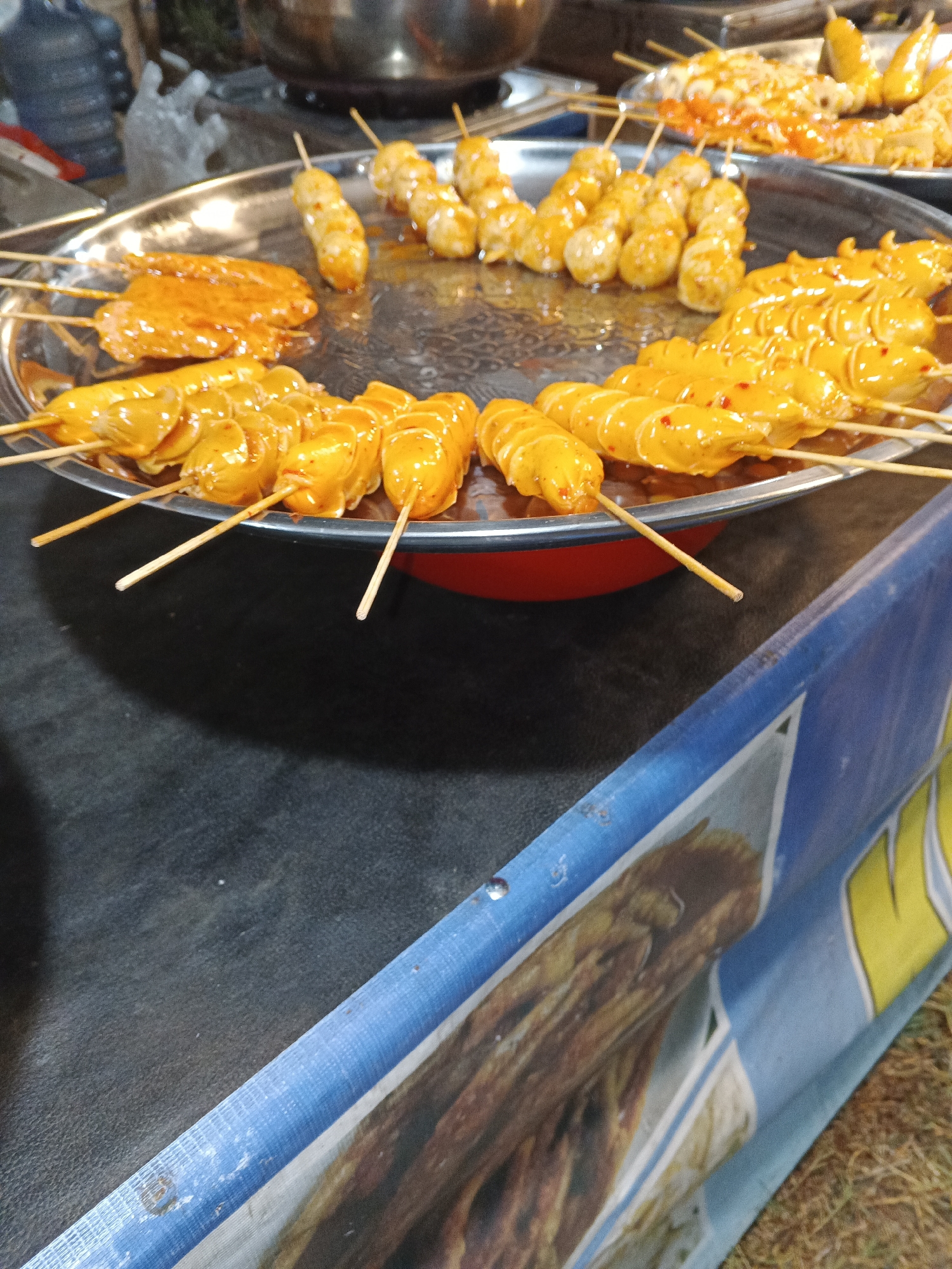 This image depicts a close-up view of various skewers of food, likely at a street food stall in Sukabumi, Indonesia. The food items, predominantly yellowish-orange in color and coated in a sauce, are arranged on wooden skewers. In the foreground and mid-ground, several skewers of what appear to be sausages or meatballs are visible, some with visible red chili flakes. A second tray or platter, further back, holds more skewers of similar food. The items are presented on a large, circular metal platter, which is partially obscuring a red bowl beneath it. The background is blurred, showing indistinct shapes that could be other food items, cooking equipment, or customers. The scene is set against a dark surface, likely a table or counter, and a blue and white banner with a printed image of food is visible in the lower right portion of the frame. The lighting suggests an outdoor or semi-outdoor setting, possibly during the evening or under artificial light.
