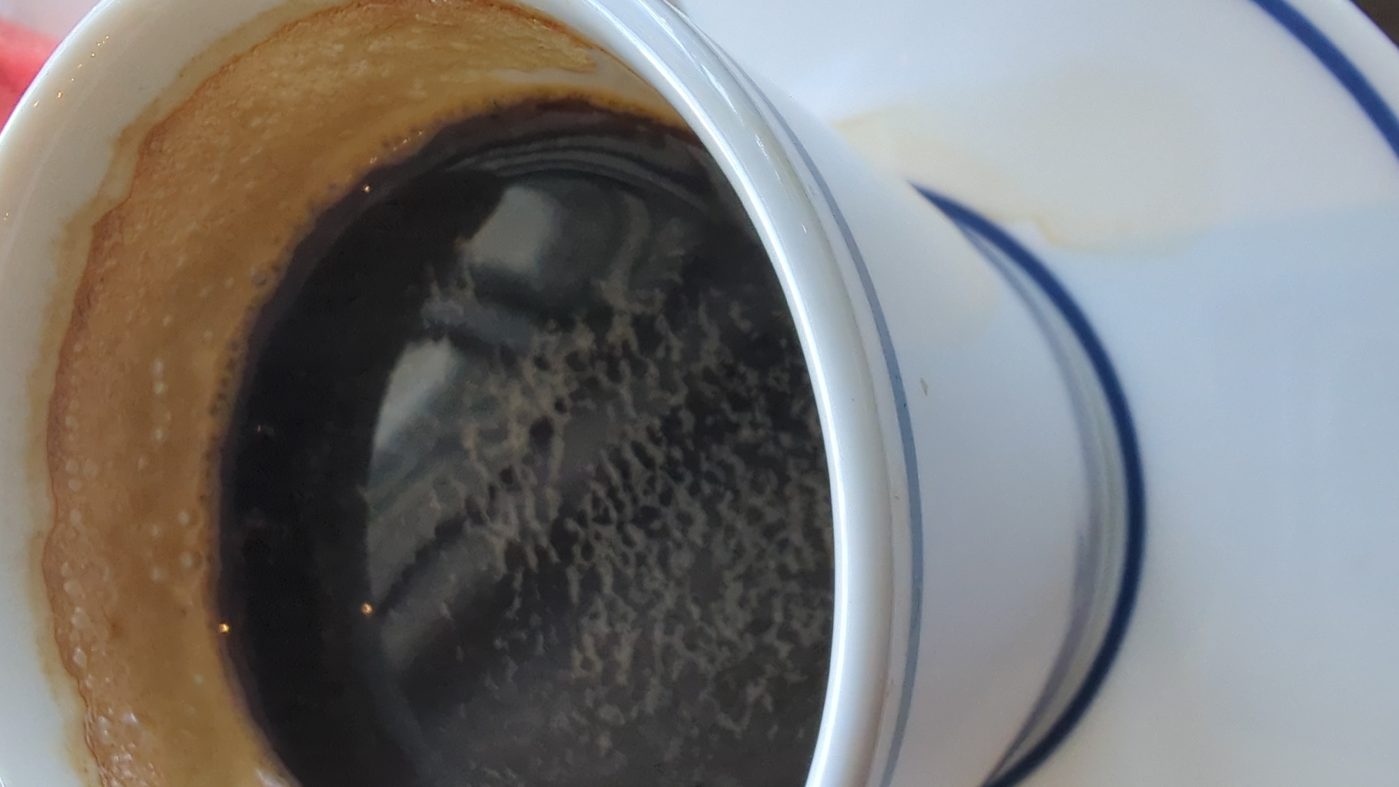 A close-up shot focuses on a white mug filled with dark coffee, topped with a thin layer of foamy crema. The mug has a subtle blue stripe along its rim and sits on a matching saucer, which also shows a faint coffee stain. The background is out of focus, but hints at a light-colored surface. The image is an interior shot, likely taken indoors. No specific location in Larkollen, Norway, can be identified from the image.