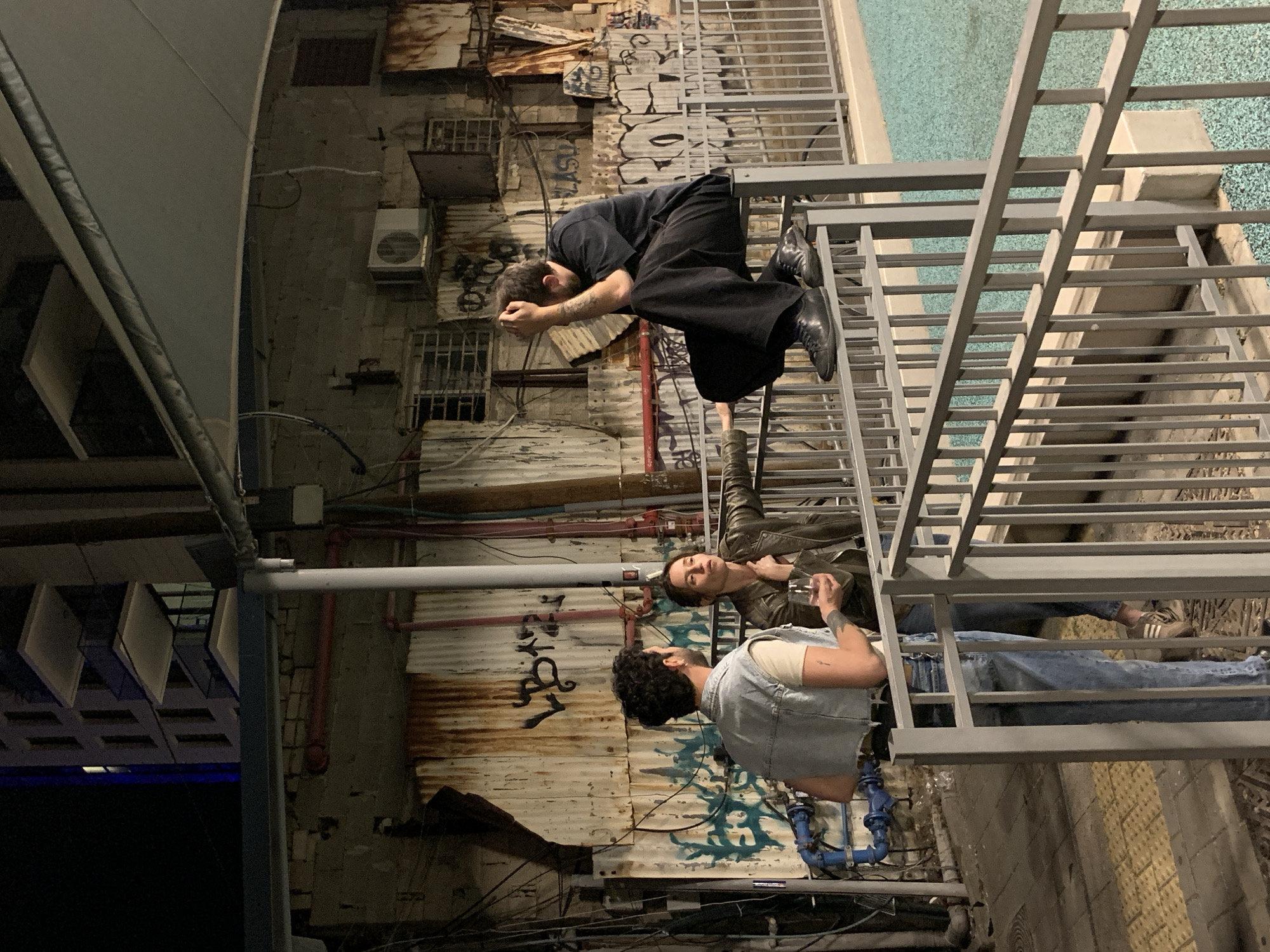 A nighttime outdoor scene features three individuals interacting on a metal staircase structure against a backdrop of weathered buildings with graffiti and various architectural elements. The location is plausibly Tel Aviv, Israel, given the urban environment. The individual closest to the top of the image is bent over the railing. He is wearing a black outfit, boots, and has dark hair with a beard. Below him is another person leaning against the railing, wearing a jacket, holding what appears to be a glass and wearing shoes. The last person is wearing a light-colored t-shirt, jeans, and a denim vest and is standing on the staircase. Graffiti and pipeworks are visible on the walls of the surrounding buildings.