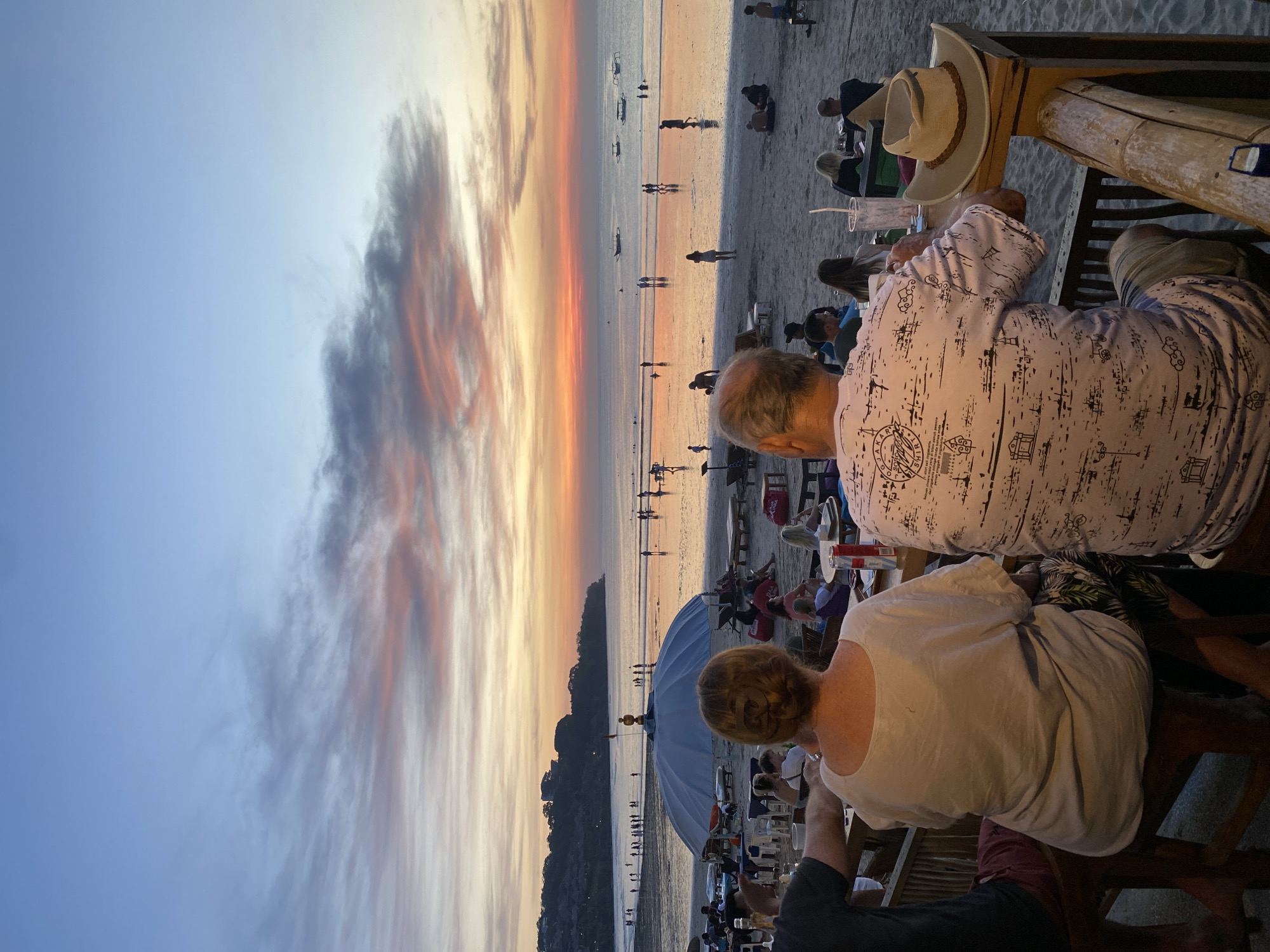 A sunset view from a beach bar in Kuta Selatan, Indonesia. Patrons are seated at a table with a straw hat hanging from a nearby post. The beach is populated with people. The sky displays colors of orange and blue.