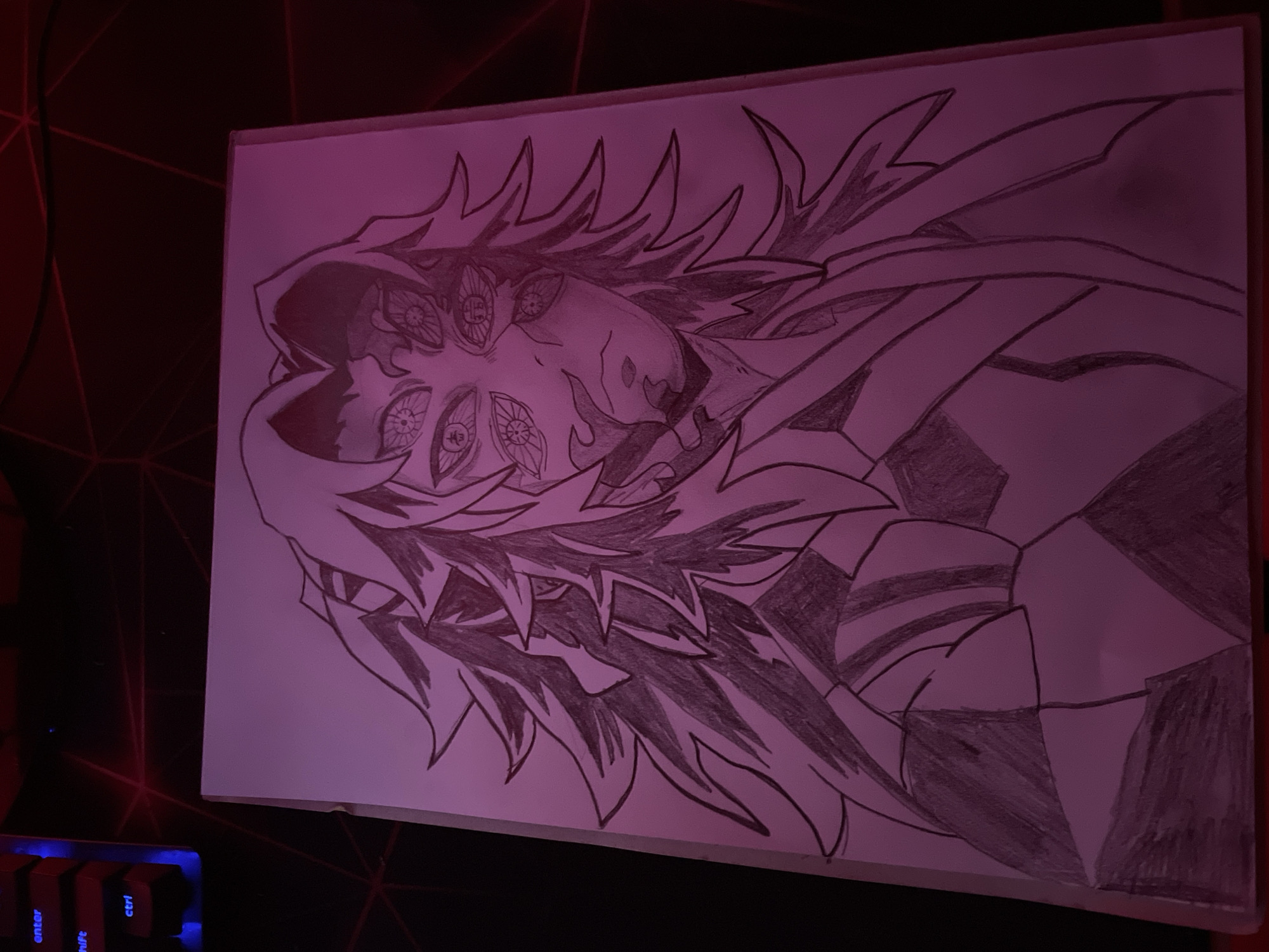 The image is a photograph of a drawing of a character with multiple eyes, spiky hair, and geometric patterns on their clothing. The drawing is on a sheet of white paper. The paper is placed on a dark surface, possibly a desk or table, with a keyboard visible in the lower left corner. The lighting in the room is dim, with a red or purple hue. The photograph appears to be taken at an angle. There is no indication the image was taken in Zlatograd, Bulgaria.