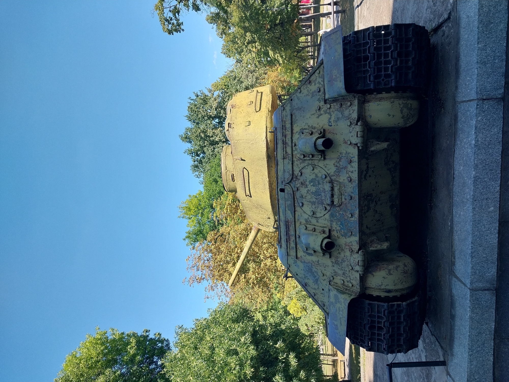 The image shows a Soviet-era T-34 tank on display, likely as a monument, in a park setting. The tank is painted a faded olive green with visible signs of wear and tear, including rust and peeling paint. It is positioned on a stone pedestal, with its tracks resting on the stone base. Behind the tank, a clear blue sky is visible above a variety of trees, some with green foliage and others with autumnal yellow leaves. In the background, there are hints of a park walkway with benches and some distant figures, suggesting this is a public space. The overall impression is of a historical artifact situated in a serene outdoor environment. This specific tank, a T-34-85 model, is located at the WWII Museum in Dnipro, Ukraine, commemorating the Second World War.