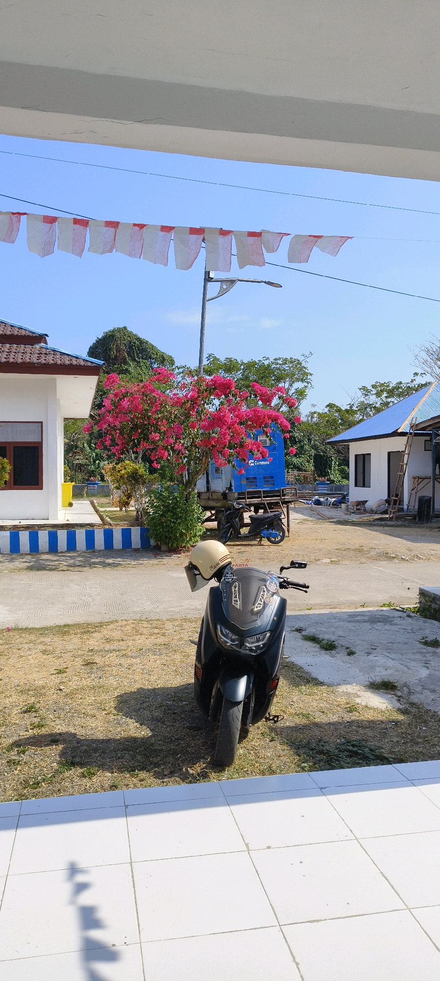 In Waisai, Indonesia, a dark grey scooter is parked on a paved area in front of a building. A light-colored helmet rests on the scooter's handlebars. Behind the scooter, a vibrant pink bougainvillea tree blooms in front of a blue generator. To the left, a white building with a brown tiled roof is visible, and to the right, another white building with a blue roof has a ladder leaning against its side. Red and white flags are strung across the top of the image, suggesting a national celebration. The sky is clear and blue, indicating a sunny day.