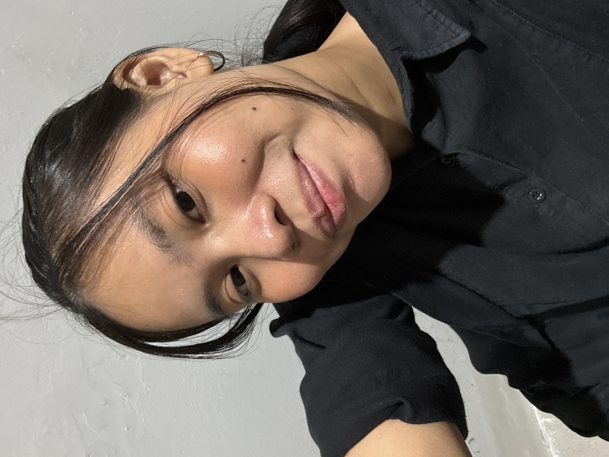A young woman with dark hair pulled back in a ponytail and a few loose strands framing her face, is captured in a close-up, head-and-shoulders selfie. She is wearing a black collared shirt. The background consists of a textured, light-colored wall. The lighting in the image is artificial, creating highlights on her forehead and cheeks, suggesting a slightly oily complexion. She is looking directly at the camera with a slight smile. The photo appears to have been taken indoors, in a residential setting.