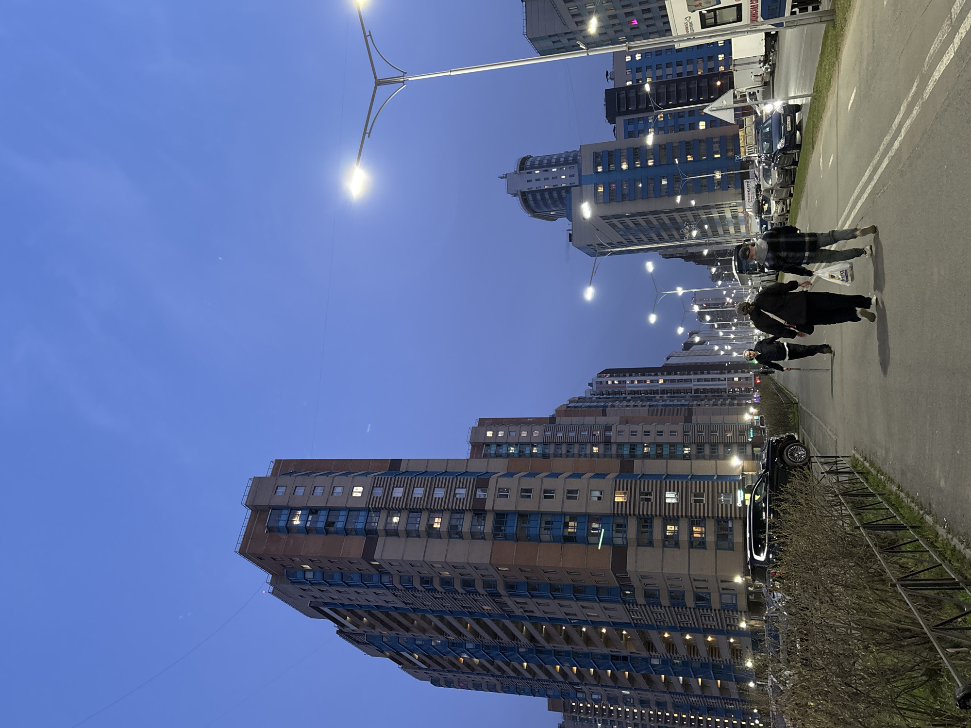 The image depicts a street in the city of Kudrovo, Russia, at twilight. Several tall apartment buildings are visible, their windows lit. Streetlights illuminate the scene. A paved road runs along the right side of the frame, with a metal railing separating it from a grassy area. People are walking along the road. Cars are parked along the side of the road.