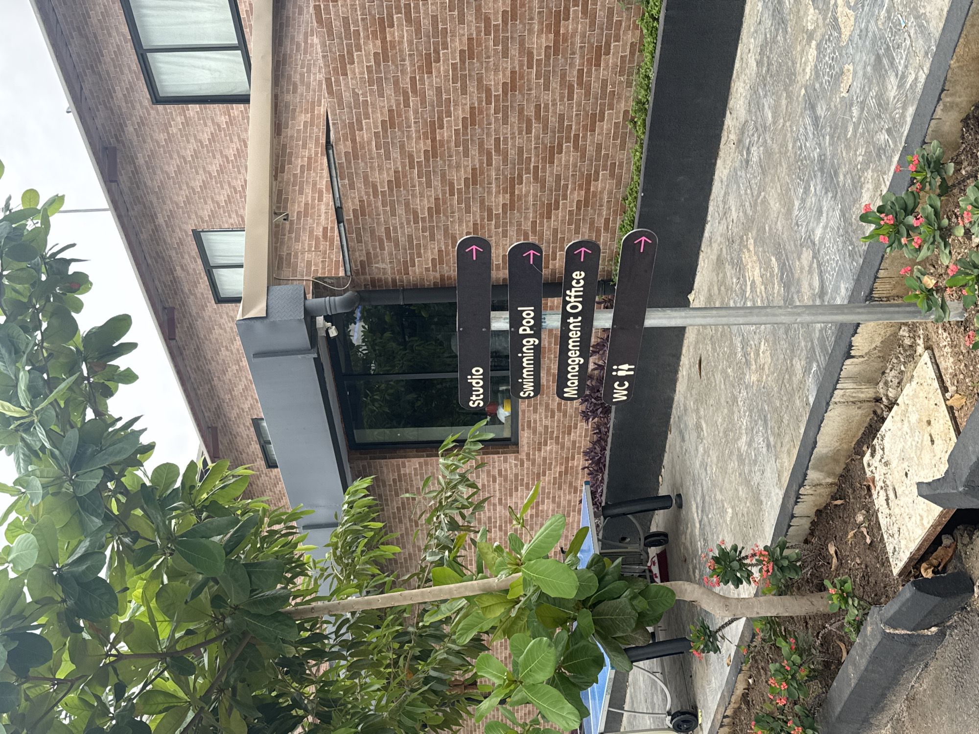 The image shows a signpost with directions to Studio, Swimming Pool, Management Office, and WC (toilet). The signpost is located near a brick building with windows. There is a tree with green leaves in the foreground and a paved walkway alongside the building. The image likely depicts an outdoor area within a building complex, with directional signage for various facilities. The location is presumed to be in Abuja, Nigeria. No people are visible in the image.