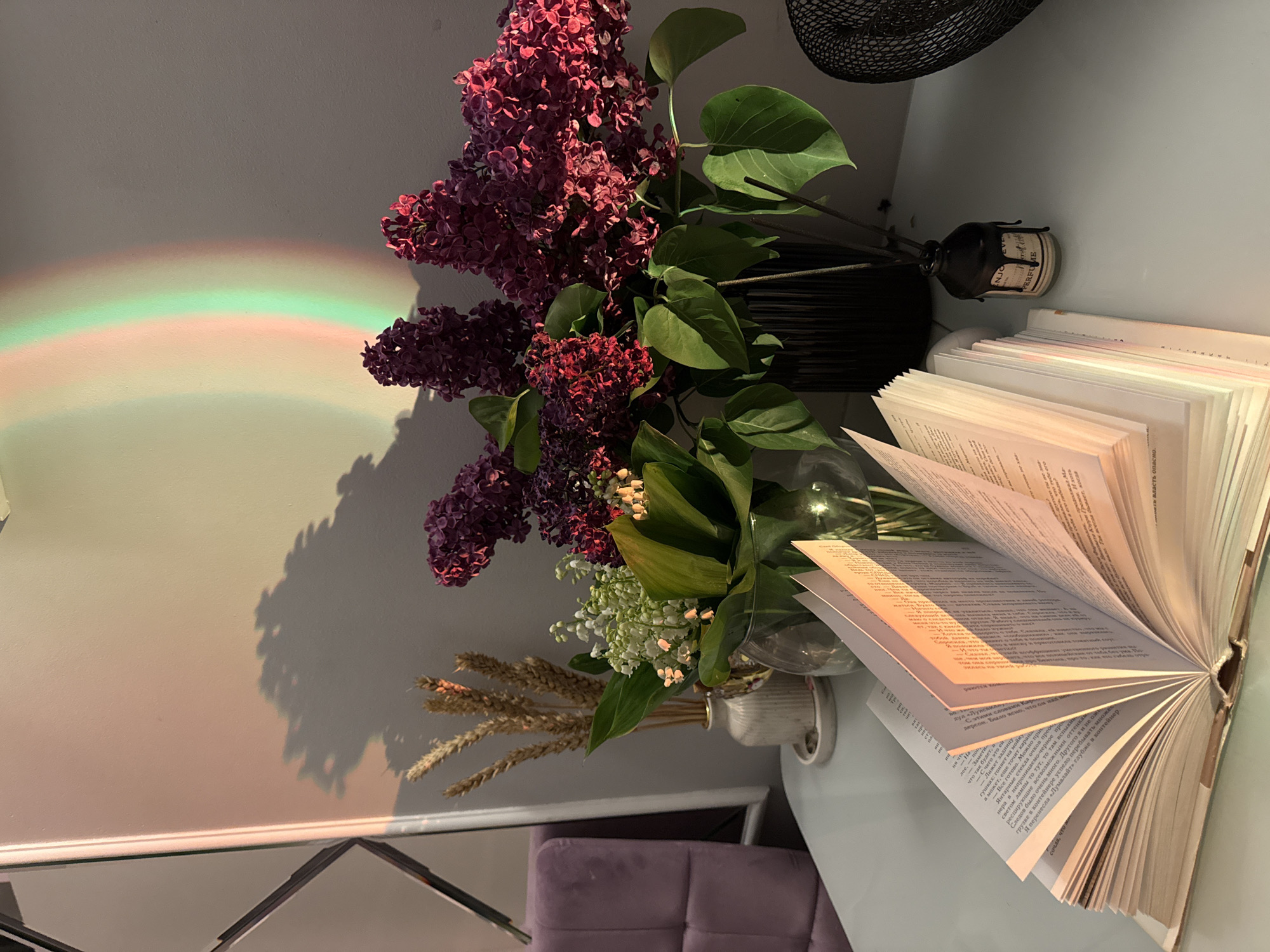 An indoor scene features a vibrant bouquet of flowers and an open book on a light-colored table, illuminated by natural light. The main subjects are large clusters of purple lilacs and smaller sprigs of white lily-of-the-valley, arranged in different vases. A dark, ribbed vase holds the lilacs, while a smaller white vase contains the lily-of-the-valley and dried wheat stalks. An open book with numerous pages fanned out rests on the table, with text visible on its pages. Beside the book, a reed diffuser bottle, dark-colored with a cream label, can be seen.
The setting appears to be a cozy room, with a light-colored wall serving as the background. A distinctive rainbow-colored streak of light, ranging from green to pink, is cast horizontally across the wall, indicating natural light filtering through a prism or glass object. Shadows of the flowers and leaves are prominently displayed on the wall, suggesting direct sunlight. Part of a dark-colored fan is visible at the top edge of the image. In the lower left corner, a partial view of a purple upholstered sofa or cushion and a reflective surface (possibly a mirror or table edge) can be seen.
The scene conveys a tranquil atmosphere, possibly a moment of quiet reading or contemplation. The visible text on the book pages includes snippets of what appears to be Russian, such as "С этими словами Кари" (With these words Kari), "Было ясно" (It was clear), and "Я перенесла «Лумалайт» глубже в контейнер" (I moved "Lumalight" deeper into the container). On the reed diffuser bottle, the words "ENJOY" and "PERFUME" are visible. The time of day is likely late morning or afternoon, given the sunlight and shadows.