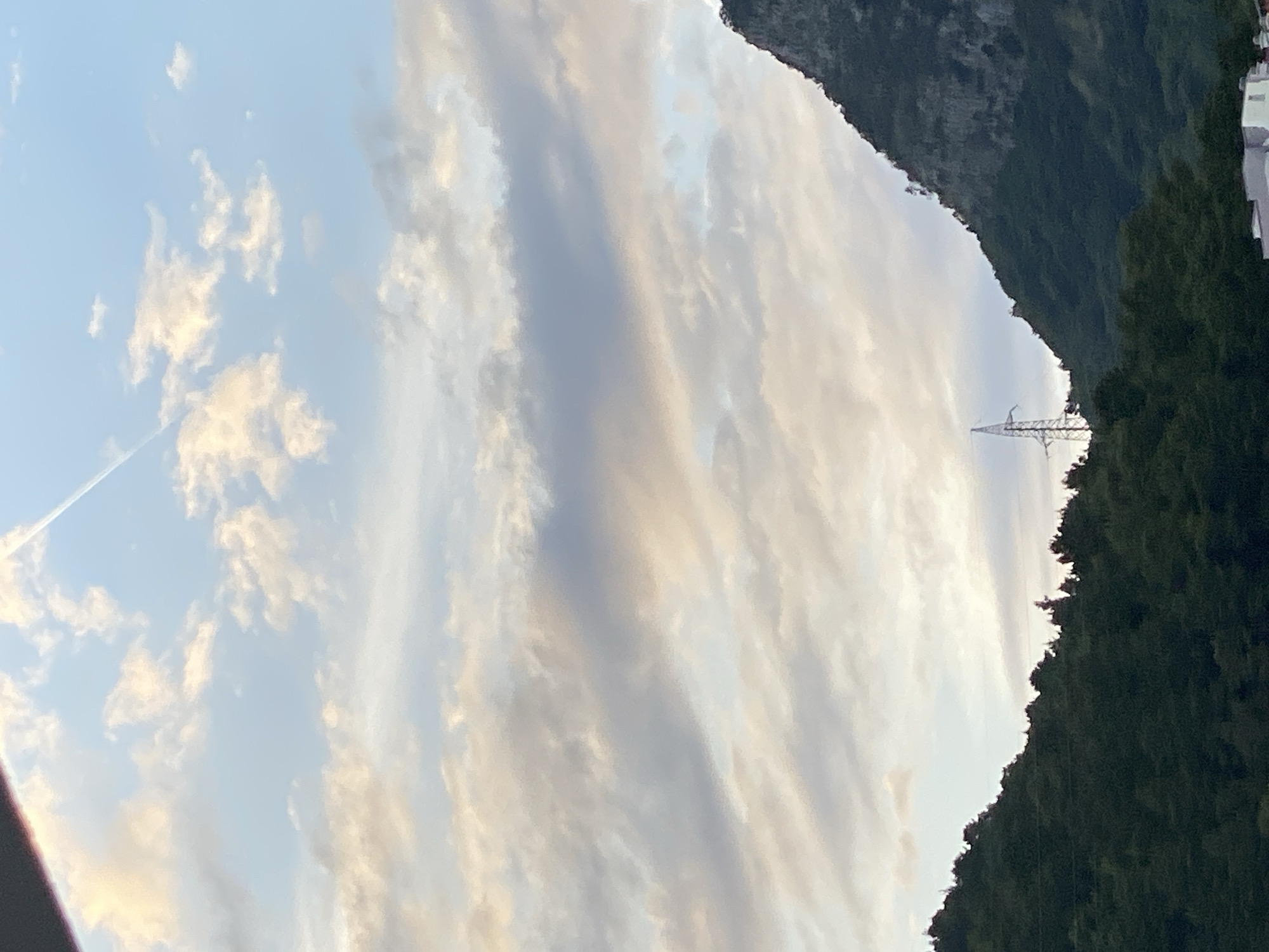 The image displays a mountainous landscape under a partially cloudy sky, located in Budva, Montenegro. The upper sky is light blue with scattered white, wispy clouds, one of which is traversed diagonally from upper left to upper right by a thin, white aircraft contrail. The middle and lower sky are dominated by larger, stratified clouds, exhibiting various shades of white, grey, and subtle light orange/yellow hues, suggesting either dawn or dusk.
In the lower left quadrant, a densely forested mountain or hill slopes upwards, characterized by dark green foliage forming a distinct silhouette against the lighter sky. On the right side, a rugged, steep mountain face rises, covered with sporadic vegetation and exposed rock formations, particularly in its upper visible sections.
Centered in the valley between these two landforms, a prominent grey metal lattice power pylon stands, its structure clearly visible against the distant landscape. At the very bottom edge of the image, the tops of two light-colored buildings are partially visible, indicating human settlement at the base of the mountains. No individuals are present in the frame.
