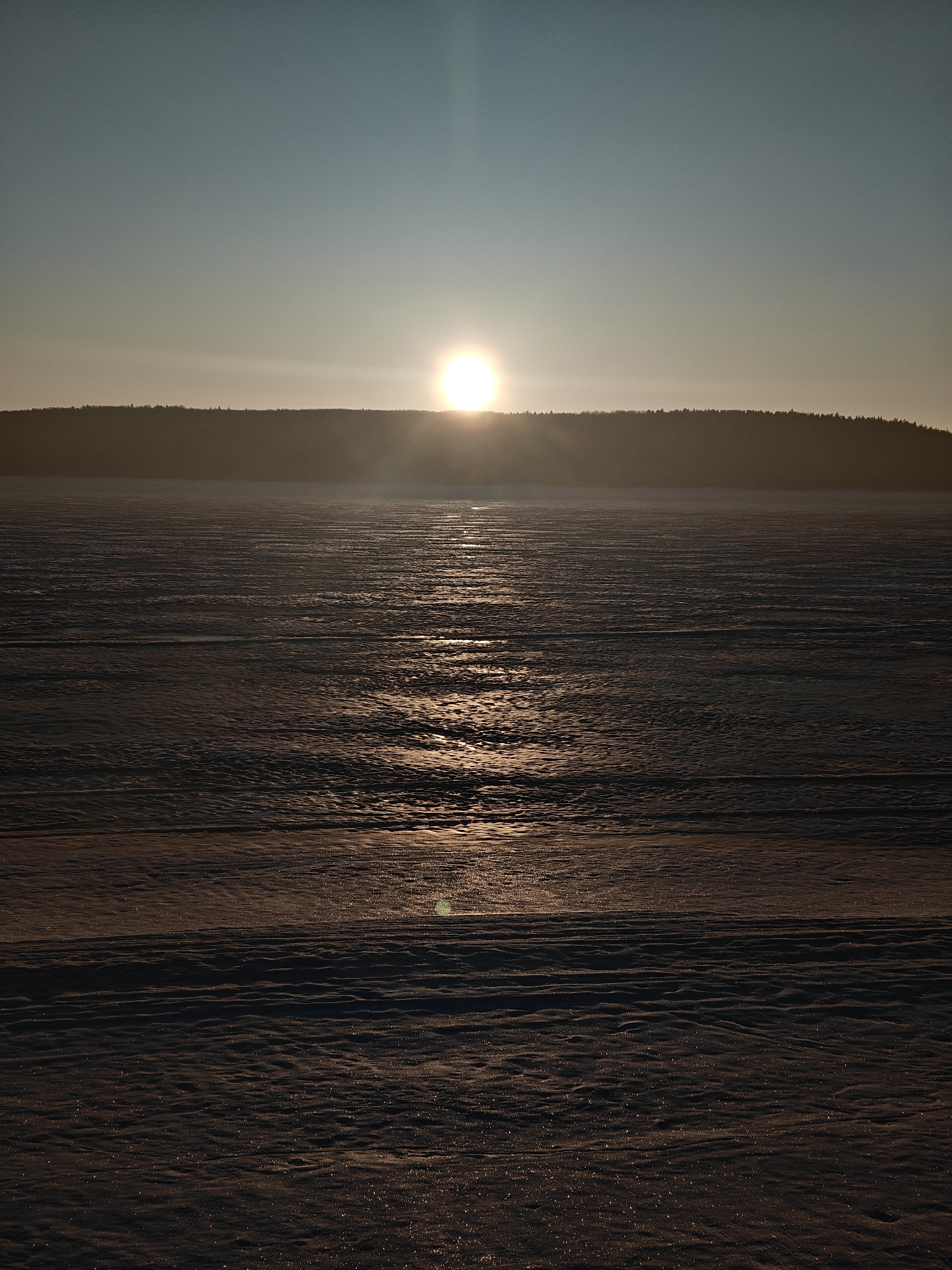 The image depicts a sunset over a snowy expanse, likely a frozen body of water, with a treeline visible in the distance. The sun is positioned on the horizon, casting a strong glare and light reflection across the snow. The scene is likely to take place in a northern latitude and location is in the city of Chaikovsky, Russia. There are no people visible in the image. The environment suggests a cold climate, with the snow covering the landscape. The actions are limited to the setting sun and the reflection on the snow. There are no clear interactions or additional subjects or objects beyond the described elements.