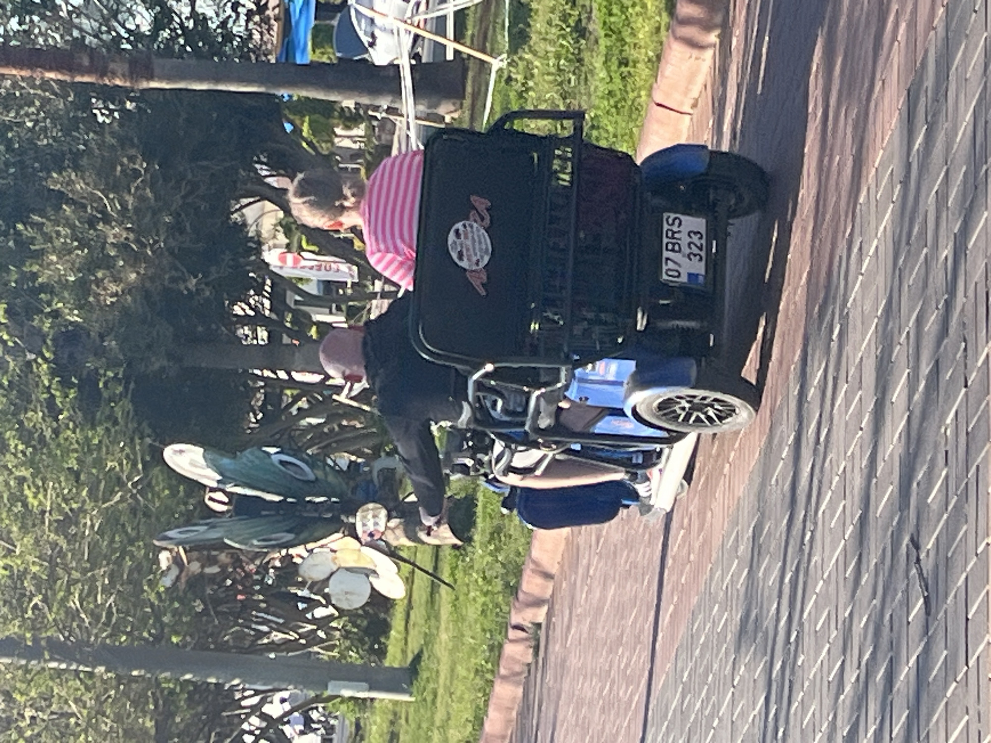 This image captures an outdoor scene in Alanya, Turkey, featuring two people on a blue motorized scooter during daylight hours. A bald or short-haired man, wearing a dark top, is operating the scooter. Behind him, a person with light hair, dressed in a pink and white striped shirt, is seated. A third individual, wearing blue shorts or pants and white sneakers, is partially visible in the lower right foreground, possibly a passenger or nearby.
The motorized scooter is blue with a black rear storage box or seat back that prominently displays "MONZA" in pink text along with a circular emblem. The scooter's license plate, located at the rear, clearly reads "07 BRS 323," with the "07" prefix confirming its registration in Antalya Province, which includes Alanya.
The scooter is situated on a paved walkway constructed from reddish-brown and gray interlocking bricks, with green grass and numerous trees lining the left side of the path. A striking feature in the background is a large, decorative flower-like sculpture or wind spinner, composed of white and teal/green metallic petals, positioned among other garden decorations. In the distance, through the foliage, glimpses of light-colored structures, possibly market stalls or tents, and a partially obscured sign with red lettering can be seen.
The scene appears to be set on a bright, sunny day, evidenced by the clear illumination and visible shadows on the pavement, suggesting good weather conditions for an outdoor excursion.