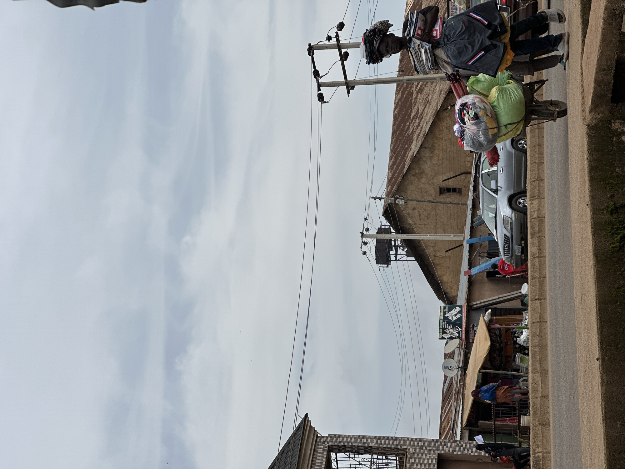 In the city of Bukuru, Nigeria, a person is pushing a wheelbarrow laden with various items, including bundles of folded clothes and bags filled with merchandise. The person is walking on a paved road. They are carrying more folded clothes stacked on their head and are holding a few items in their hands. Behind them, a silver sedan is parked on the side of the road. Further back, a building with a corrugated iron roof stands, and to its left, a smaller structure with a thatched roof serves as a shop. The shop has merchandise displayed outside, including bottles, bags, and other goods. A sign advertising "FANCY HAIR DRESSING ABILITY BARBAS" is visible on the building. The sky is overcast, with scattered clouds. In the foreground, a concrete barrier separates the road from a lower level. The image captures a bustling street scene with elements of commerce and daily life.