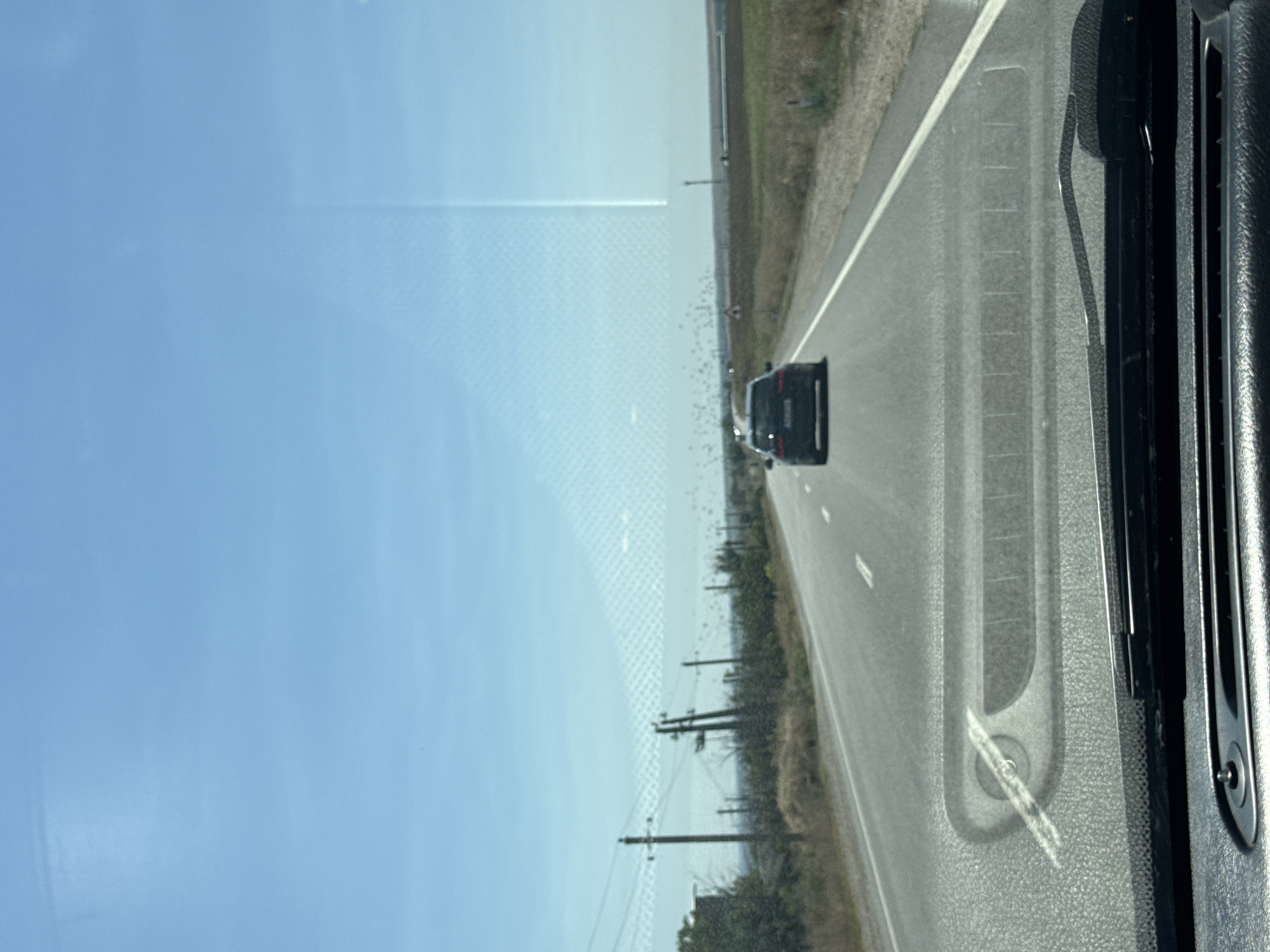 This is a rear-view image taken from inside a vehicle traveling on a paved road. The road has a white lane marker. In the distance, a black car is driving ahead. The surrounding environment consists of dry grass and some vegetation, with utility poles and wires lining the left side of the road. The sky is clear and blue. The lower part of the image shows the dashboard and windshield of the vehicle. There are no people visible other than the driver implied by the perspective.