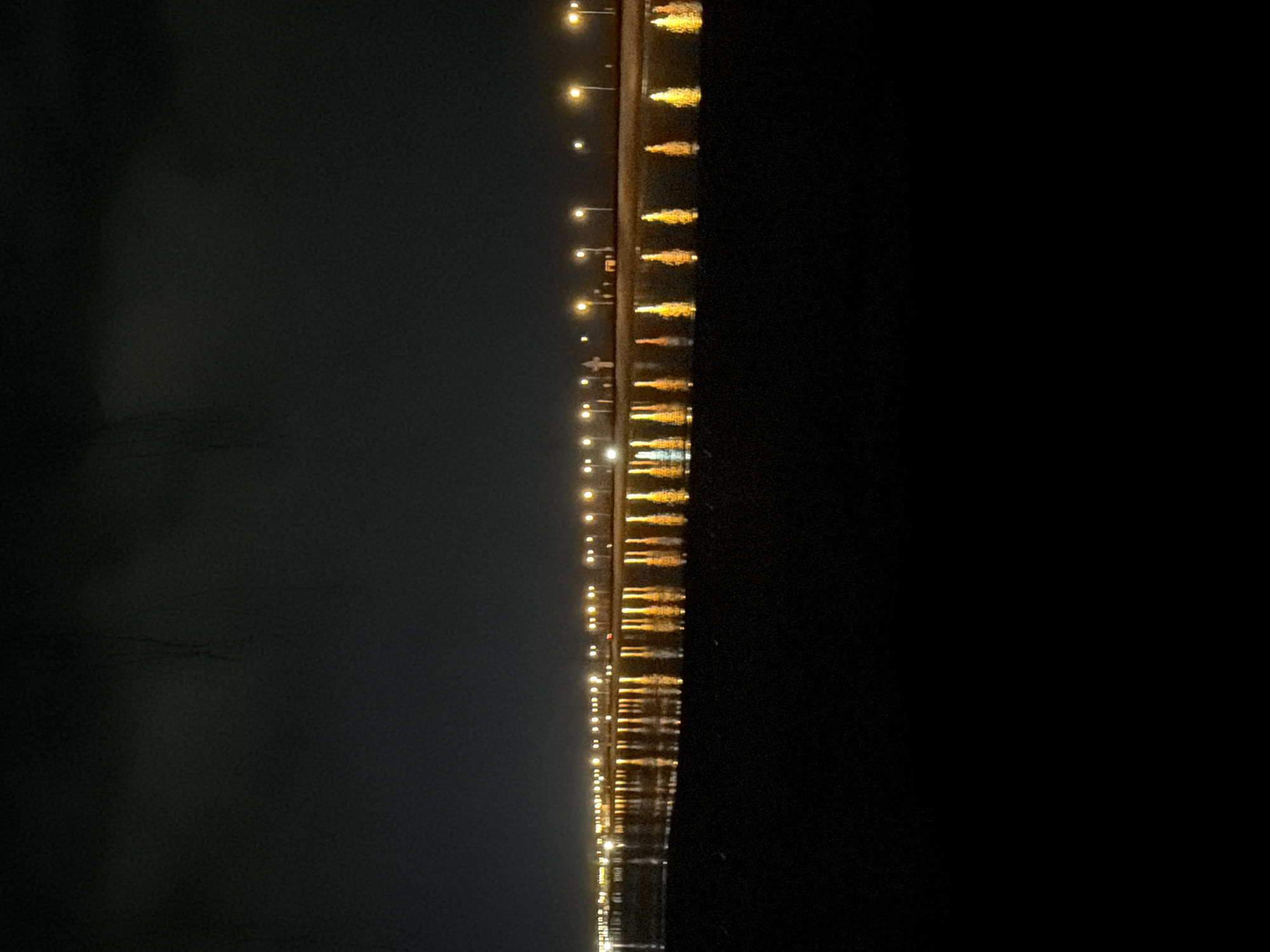 The image displays a nighttime scene, likely depicting a long bridge or pier extending horizontally across a body of water. Numerous warm-toned lights, resembling streetlights, are evenly spaced along the structure. Each light casts a distinct, shimmering reflection on the dark surface of the water below, indicating calm or lightly rippled conditions.
The left side of the frame, representing the sky or distant background, is a deep, dark grey, while the right side is completely black, suggesting the expanse of dark water or an area devoid of light sources. The focus is entirely on the illuminated structure and its reflections. There are no people or specific activities taking place. The time of day is clearly night, and the weather appears clear, allowing for sharp reflections. No visible text is present in the image. The setting conveys a sense of peaceful isolation.