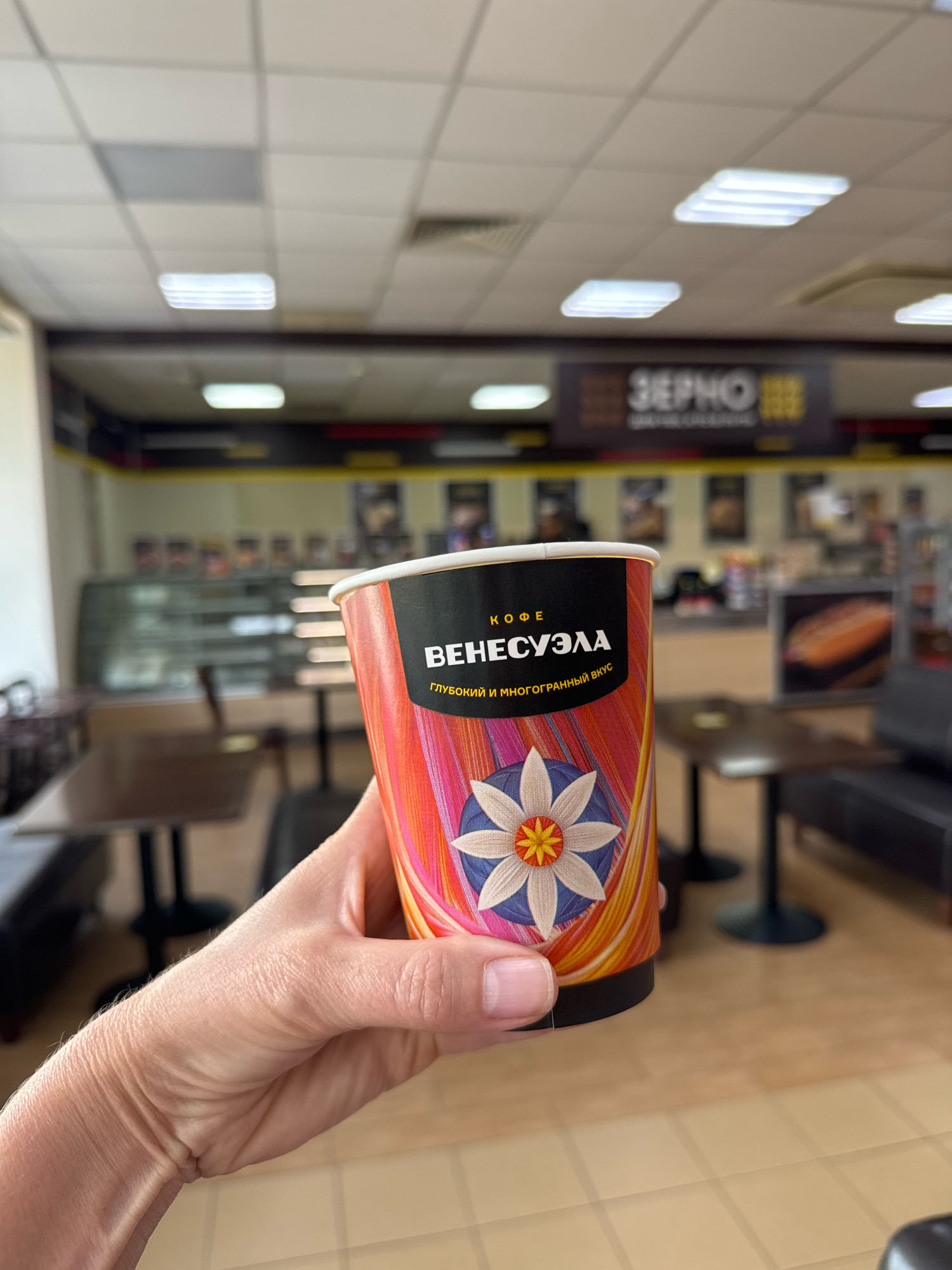 A hand holds a disposable coffee cup in the foreground, with the cup's colorful design featuring a floral pattern and text in Cyrillic. The background appears to be the interior of a cafe or convenience store, with tables, chairs, display cases, and overhead fluorescent lighting visible. A banner with Cyrillic text, "ЗЕРНО," which translates to "GRAIN," is prominent on the back wall. The overall scene suggests a casual purchase and consumption of coffee within a commercial establishment. There is no indication of the specific city of Novoselitsy, Russia, in the image itself.