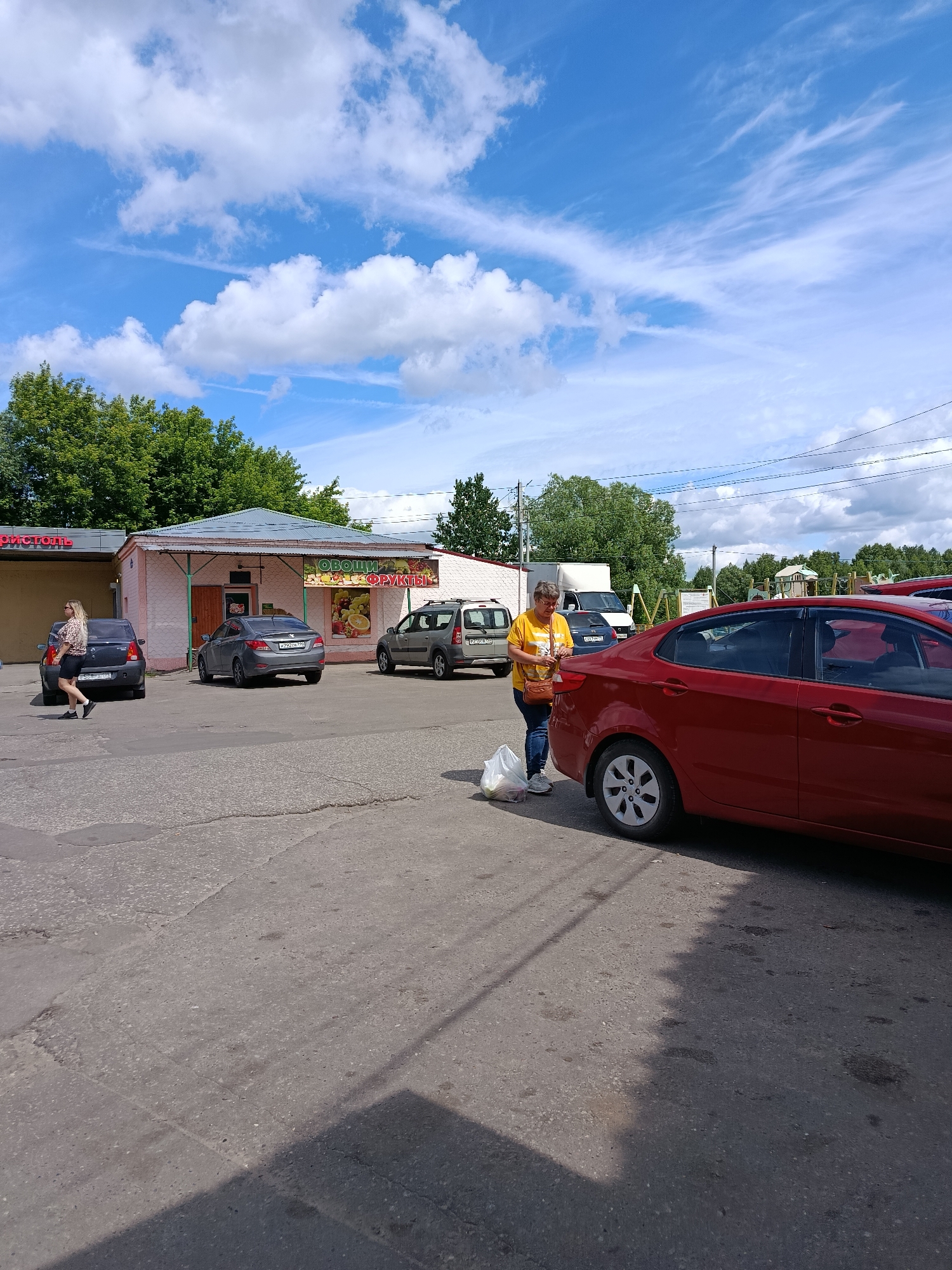 The image depicts a street scene in Veraya, Russia, on a sunny day with a partly cloudy sky. In the foreground, a red sedan is parked. To the left of the sedan, a woman wearing a yellow t-shirt and blue jeans is standing, holding a plastic bag. Behind her, a gray sedan and a dark-colored sedan are parked. In the background, a low-rise building with a light pink facade is visible, featuring signs for "ОВОЩИ" (Vegetables) and "ФРУКТЫ" (Fruits). Further to the left, another building with a red sign is partially visible. Trees line the background. The ground is paved with asphalt.