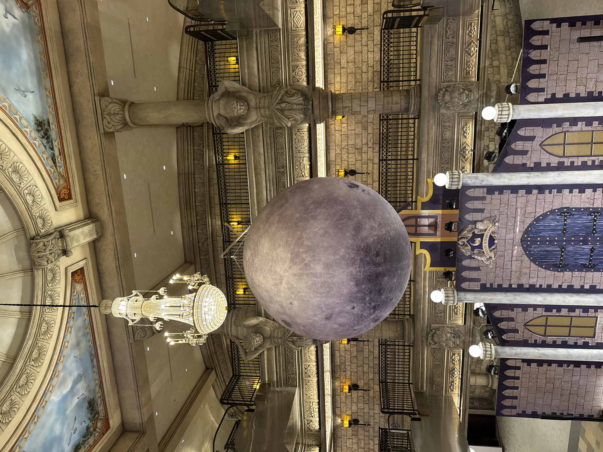 The image shows an interior scene with elaborate architectural details. A large, spherical object resembling the moon hangs from the ceiling, dominating the view. The ceiling is adorned with classical elements, including columns, statues, murals with painted skies and birds, and ornate moldings. A chandelier is visible on the left side of the frame. Balconies with wrought-iron railings line the upper portions of the walls. The building appears to simulate a castle interior, with faux stonework and simulated windows. The scene is likely located in a commercial or entertainment venue in the city of Đà Nẵng, Vietnam.
