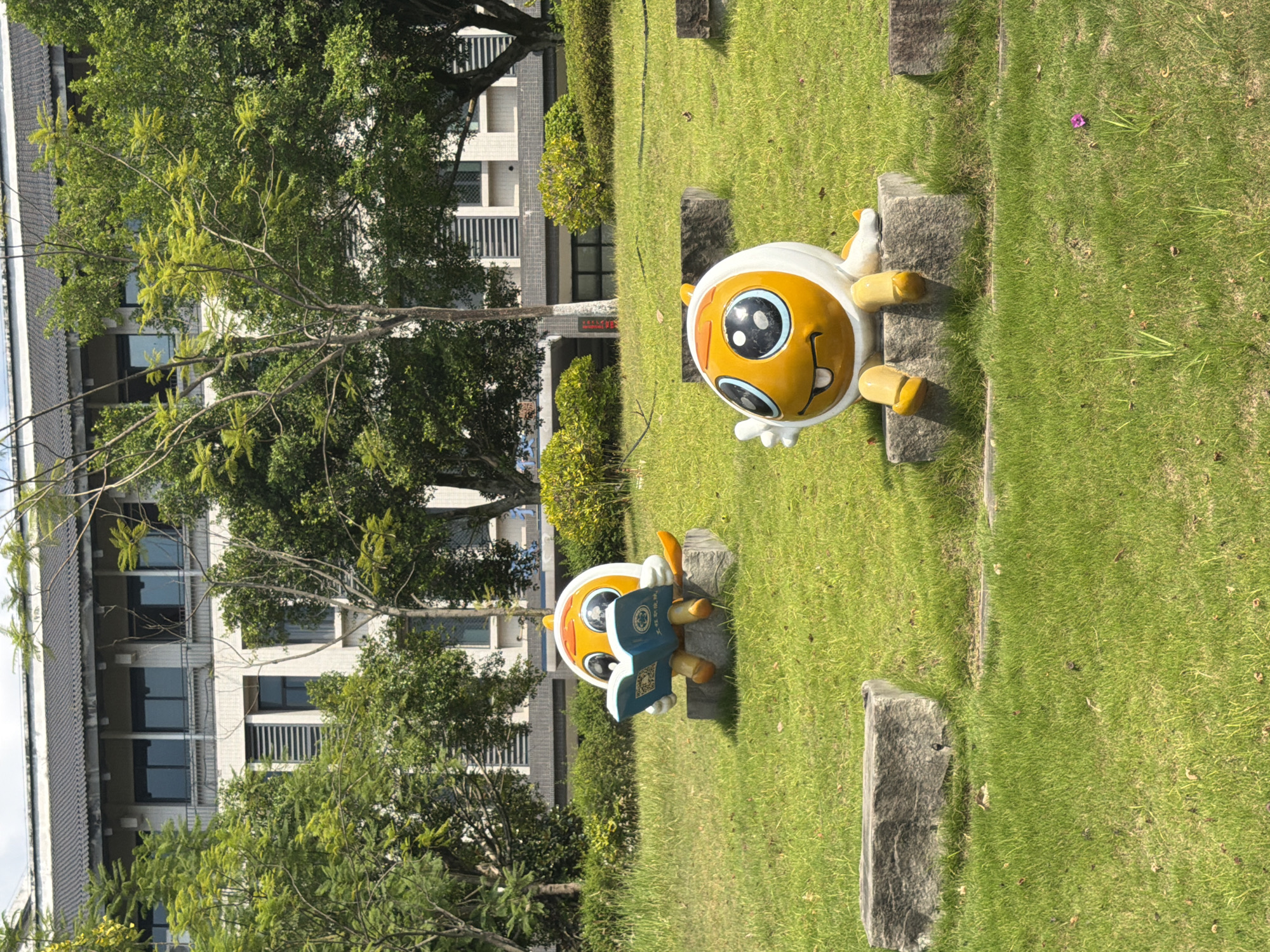 In Jiangjin, China, two cartoonish yellow and white character statues are situated on a grassy lawn in front of a modern building. The statue on the left is depicted sitting on a stone block, engrossed in reading an open blue book held in its hands. The statue on the right is also sitting on a stone block, with one hand raised in a wave and a smile on its face. The building in the background appears to be an educational institution, with multiple windows and a tiled roof. Lush green trees surround the statues and the building, suggesting a well-maintained campus environment. The lighting indicates it is daytime, with clear skies and ample sunlight.