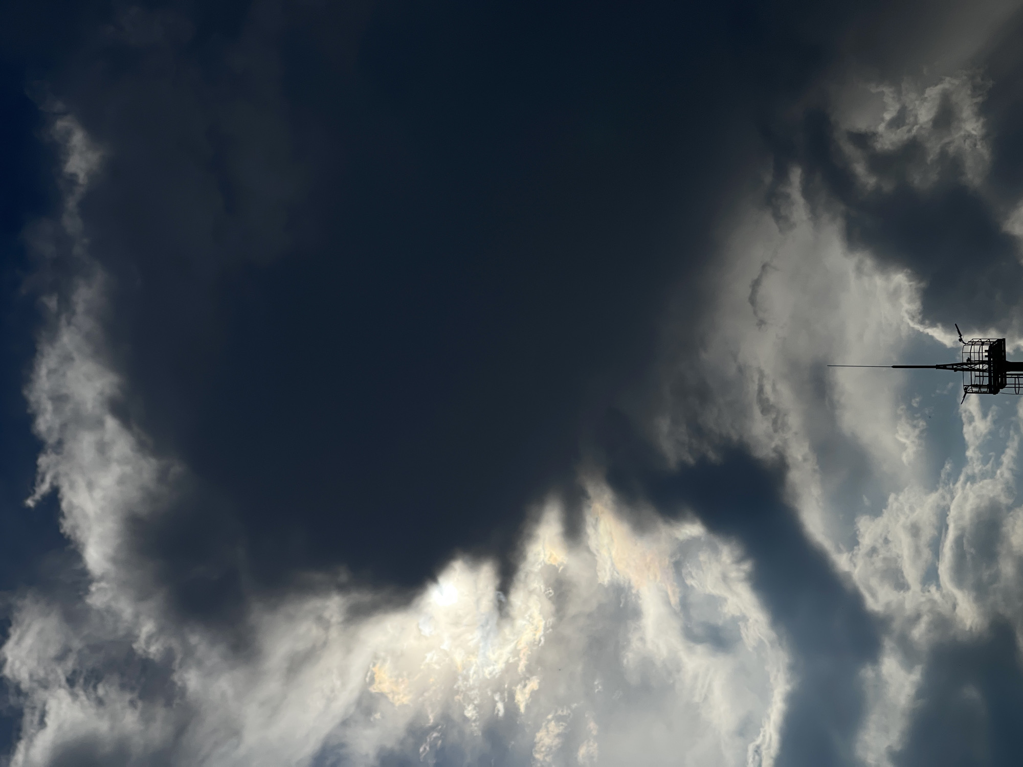 A tall, dark, and dramatic storm cloud looms over a sunlit sky, partially obscuring a bright sun. The cloud is vast, with a distinct division between the dark, stormy portion and the illuminated side. Light filters through the brighter clouds, creating an ethereal glow. In the foreground, a dark silhouette of a communication tower or similar structure pierces the bottom of the frame, its antenna reaching upwards towards the dramatic sky. The tower appears to be made of dark metal and has a platform with some equipment. The scene evokes a sense of impending weather change.