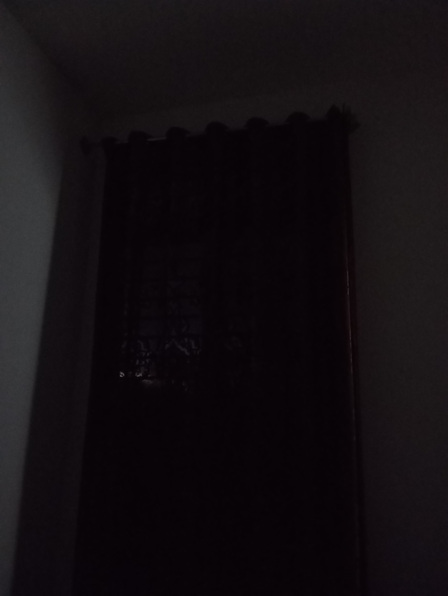 In a dimly lit room, a dark curtain is drawn across a window. The curtain appears to be made of a thick, opaque fabric, possibly velvet, with a textured surface. The top edge of the curtain is adorned with grommets or rings, suggesting it is hung from a rod. Some faint light filters through the curtain, creating a slightly lighter patch on its surface, hinting at the presence of a window behind it. The surrounding walls are dark and indistinct, with a hint of a corner visible on the left side of the frame. The overall impression is one of low light conditions, possibly evening or night. There are no people or discernible objects visible beyond the curtain and the immediate wall space.