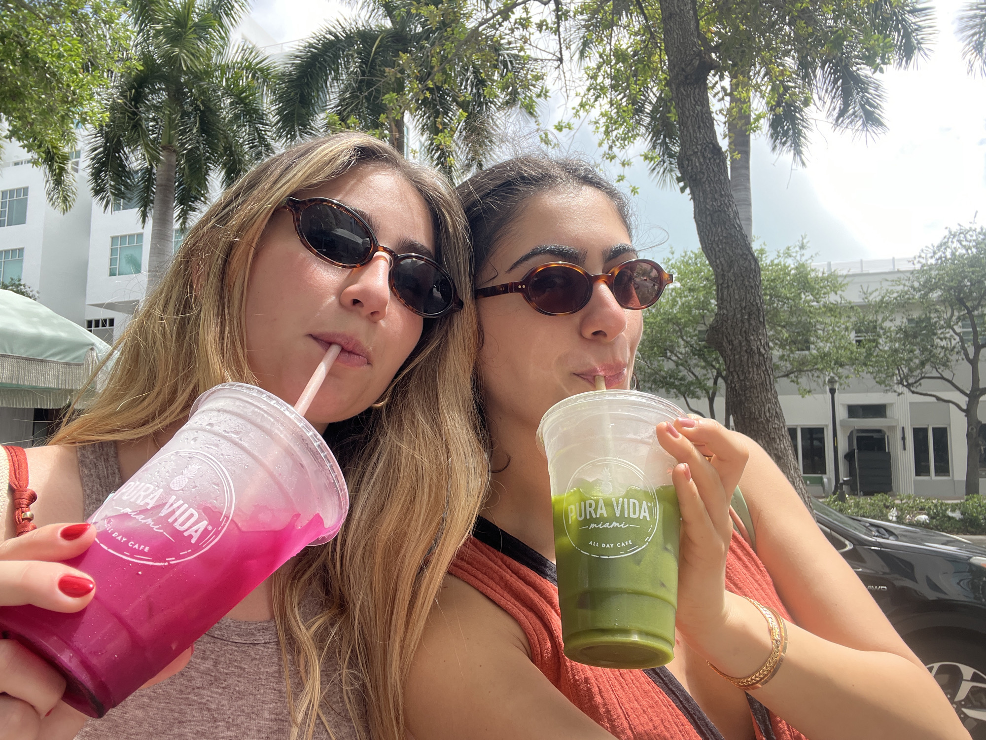 The image depicts two young women outdoors, each holding a beverage from the "PURA VIDA Miami ALL DAY CAFE". The woman on the left has long blonde hair and is wearing sunglasses and a reddish-tan tank top. She is holding a pink-colored drink. The woman on the right has dark hair pulled back, sunglasses, and an orange tank top. She is holding a green-colored drink. Both women are drinking from straws. The background shows a sunny, urban environment with trees and white buildings, indicative of a location in Miami Beach, United States of America. A dark-colored vehicle is visible in the right background.