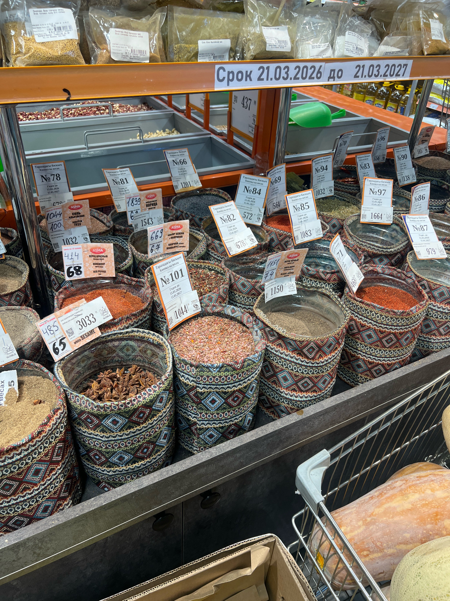 The image shows a display of bulk spices in a retail setting, likely a supermarket or market in Osh City, Kyrgyzstan. The spices are packaged in clear plastic bags and in large, decorative fabric bins. Price tags are visible, indicating product numbers and pricing in local currency. In the foreground, a shopping cart is present, containing what appear to be pumpkins and a melon. The overall environment suggests a food retail space with an emphasis on bulk goods.