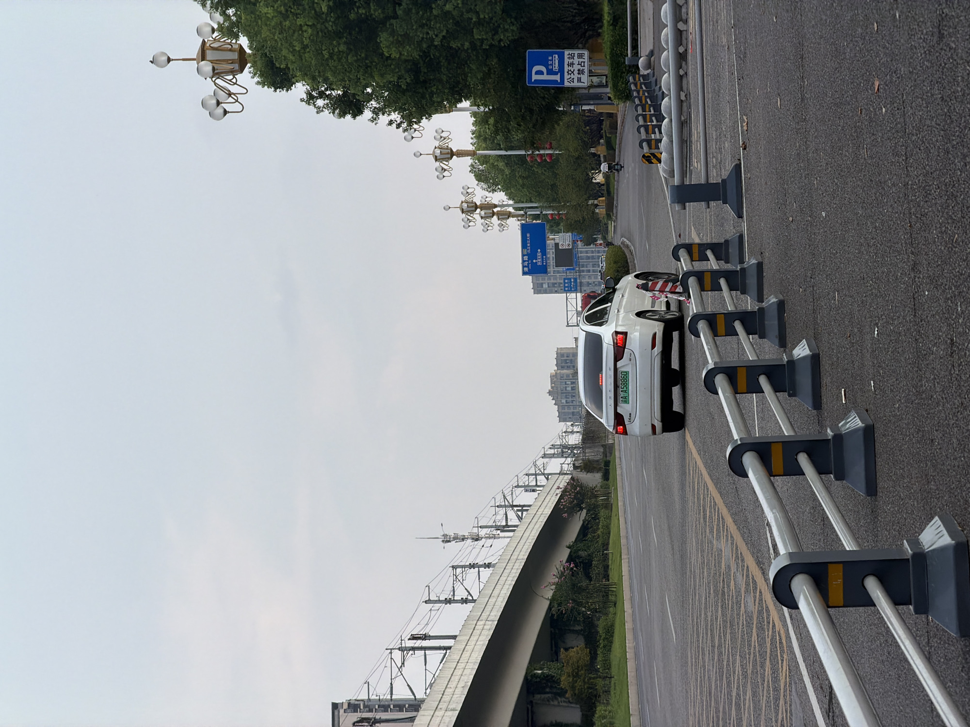 The image depicts a view of a wide, paved road in Jiangjin, China, under an overcast sky. A white sedan with license plate A A58860 is driving away from the camera on the right lane of the road. The car's brake lights are illuminated. In the foreground, a series of metal guardrails with dark grey bases and yellow markings separate the lanes of traffic. To the left of the road, a concrete overpass for an elevated railway line is visible, supported by pylons and crisscrossed with electrical wires. Greenery, including trees and bushes, lines the sides of the road and the embankment beneath the overpass. In the distance, several multi-story buildings can be seen, some with architectural details suggesting a modern urban environment. Lampposts with multiple lights are spaced along the right side of the road, and a blue "P" sign indicating parking is also present, along with Chinese text. The overall scene conveys a sense of urban infrastructure and daily transportation in Jiangjin.
