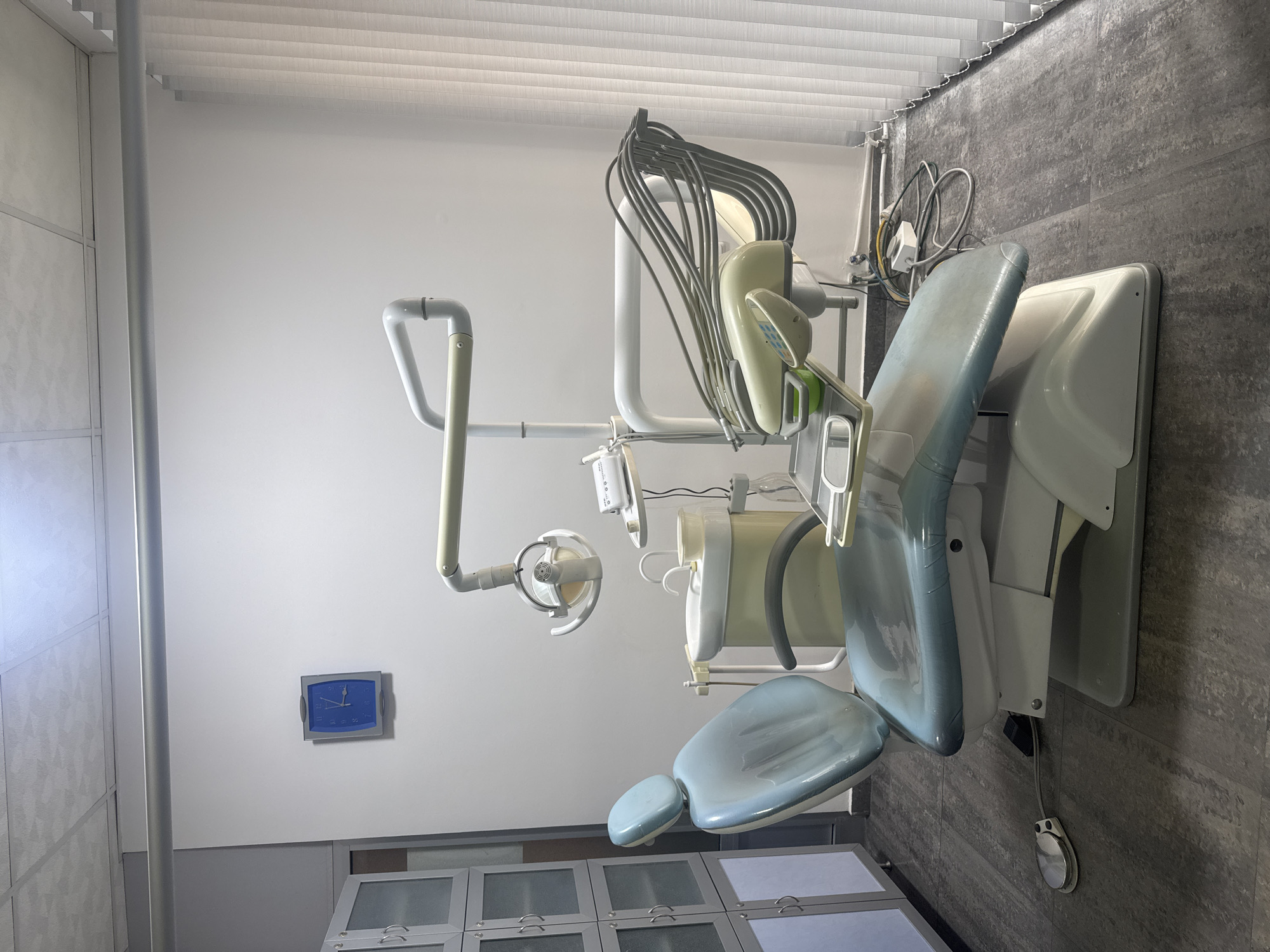 The image presents a dental examination room, likely situated within a clinic. The focal point is a modern dental chair, positioned in a neutral color scheme. The chair is surrounded by standard dental equipment, including an adjustable light fixture and various hoses and instruments typical of a dental setting. The room features light gray cabinets along the lower part of the wall and a square, blue clock hanging above them. The room's flooring appears to be textured tiles, while the upper walls are a plain white. Blinds cover a window. There are no people visible in the photograph. It is impossible to confirm the image was taken in Yerevan, Armenia, based on the visual information.