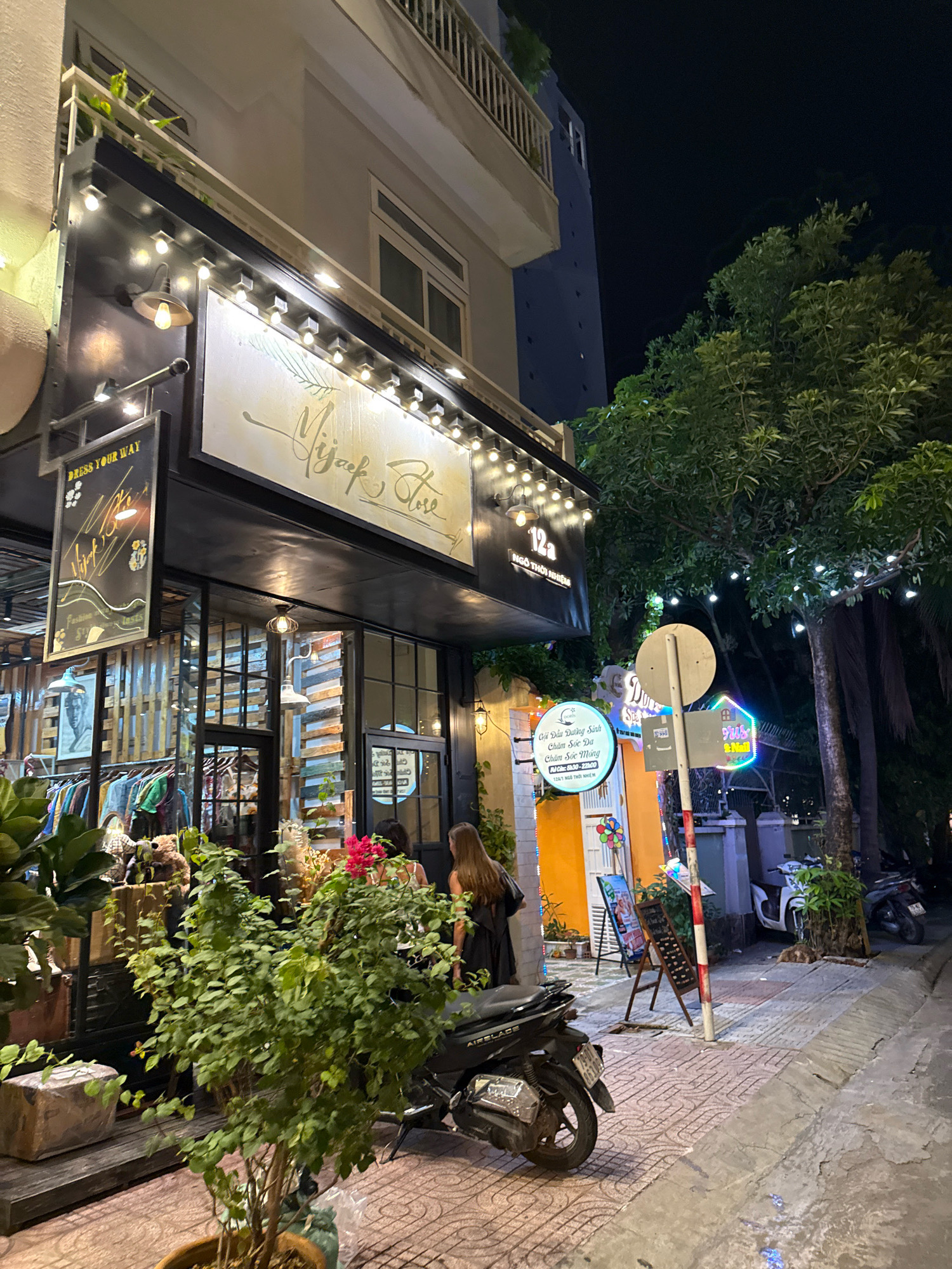 The image depicts a street scene at night in Nha Trang, Vietnam, focusing on the facade of a boutique named "Mijack Store" and its surroundings. The store features a large illuminated sign with elegant script. String lights are draped across the storefront and in the trees lining the street, creating a warm, inviting ambiance.
In front of the store, racks of clothing are visible through the glass windows, suggesting it is a clothing boutique. Several potted plants are placed outside, adding greenery to the urban setting. A scooter is parked on the sidewalk.
Two women are visible near the entrance of the store. One, with long brown hair, is wearing a black dress and appears to be walking away from the store. Another woman, also with long hair, is standing further down the sidewalk, her back to the camera, and appears to be looking at something on a display board.
Further down the street, there are other businesses with illuminated signs, including one with colorful neon lights. The sidewalk is paved with patterned tiles, and a street sign is partially visible. Palm trees and other foliage are present, characteristic of a tropical urban environment. The overall scene is one of a lively, well-lit commercial street at night.