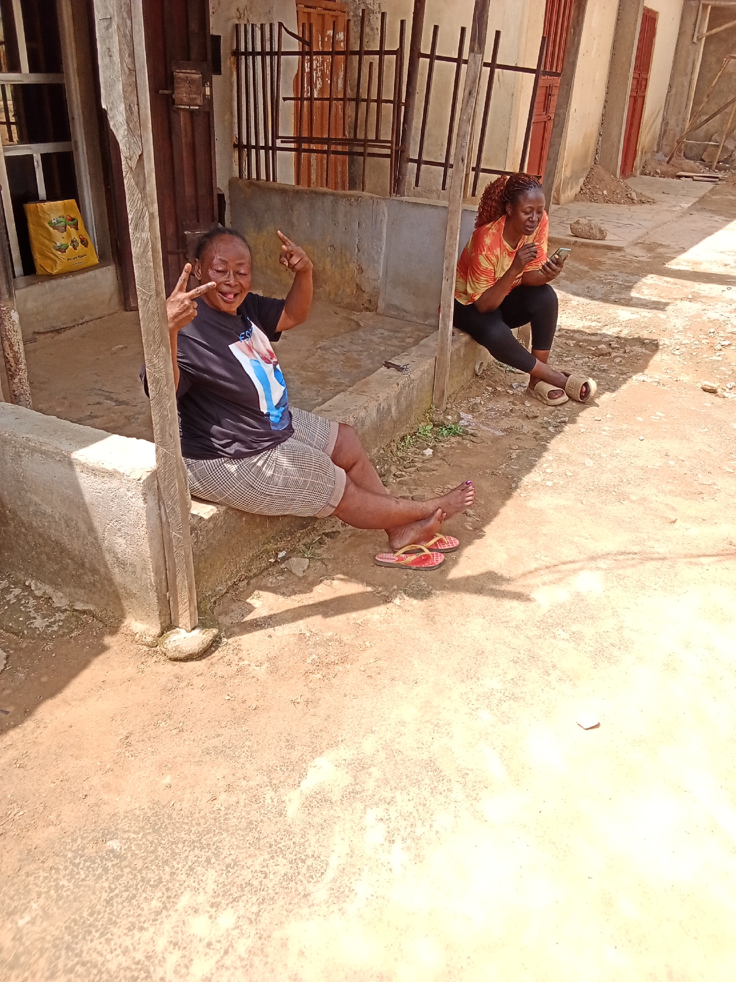 The image depicts an outdoor scene, likely in Narayi, Nigeria, with two women present. The woman on the left, wearing a black t-shirt and checkered shorts, is seated and making a peace sign with her left hand, while pointing with her right index finger. She has her eyes closed and her tongue sticking out. She is wearing red flip-flops. The woman on the right, dressed in an orange and yellow tie-dye shirt and black leggings, is seated and looking at her smartphone. She is wearing beige sandals. They are positioned near a concrete structure, with a wooden post in the foreground. The ground is sandy and dusty, with some vegetation visible. In the background, there appears to be a building with barred windows and doors. A yellow bag with graphics is placed near a window on the left side of the image. The overall impression is one of a casual outdoor gathering or break.