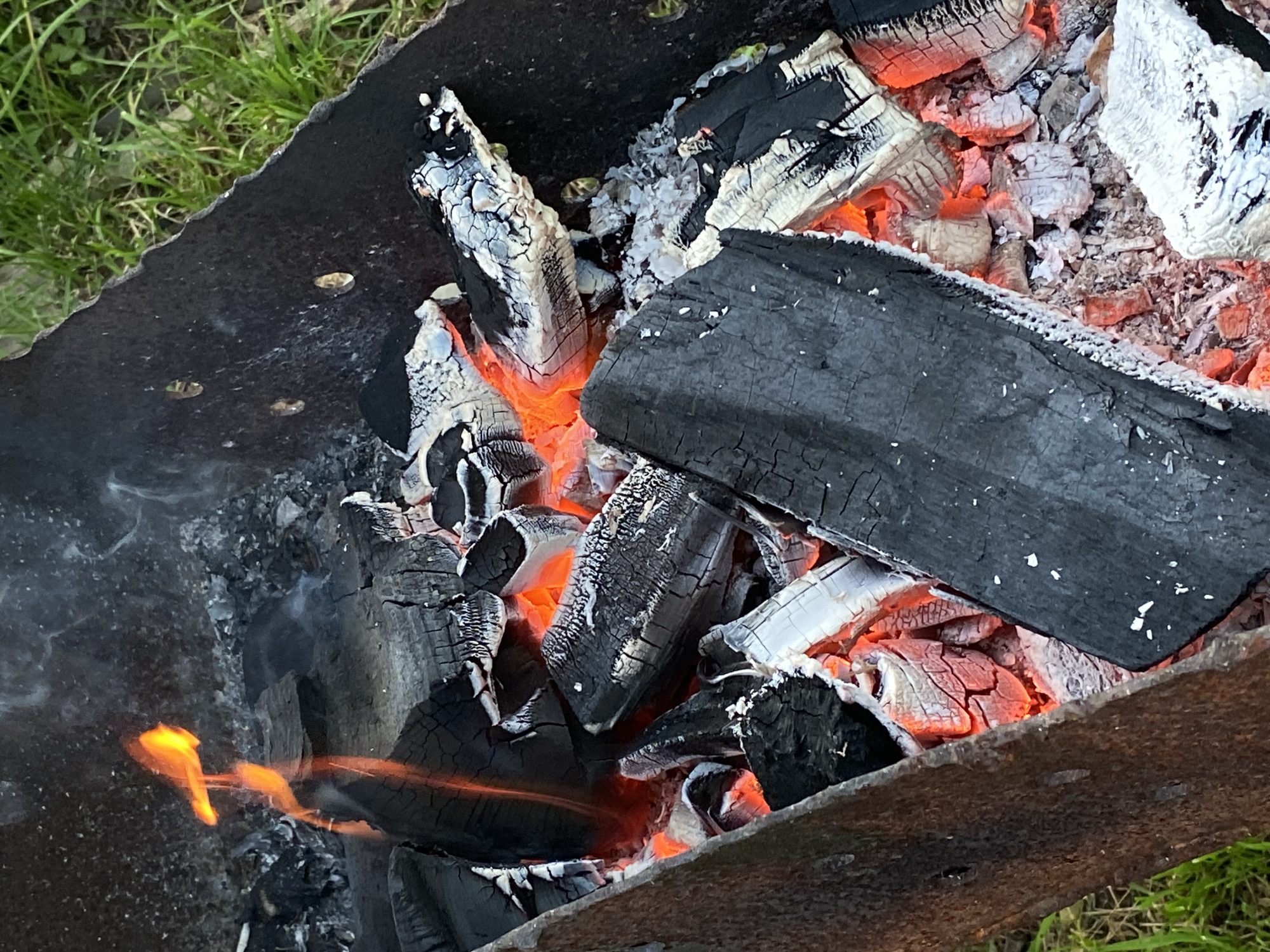 An outdoor scene captures a close-up of glowing embers and burning logs within a metal fire pit. The fire pit, appearing weathered and possibly rusted, is filled with a mix of charcoal pieces, some still actively burning with red and orange hues, while others are covered in white ash. Wisps of smoke rise from the fire. The background shows green grass, suggesting an outdoor setting. The location is identified as Novyi Ural, Russia.