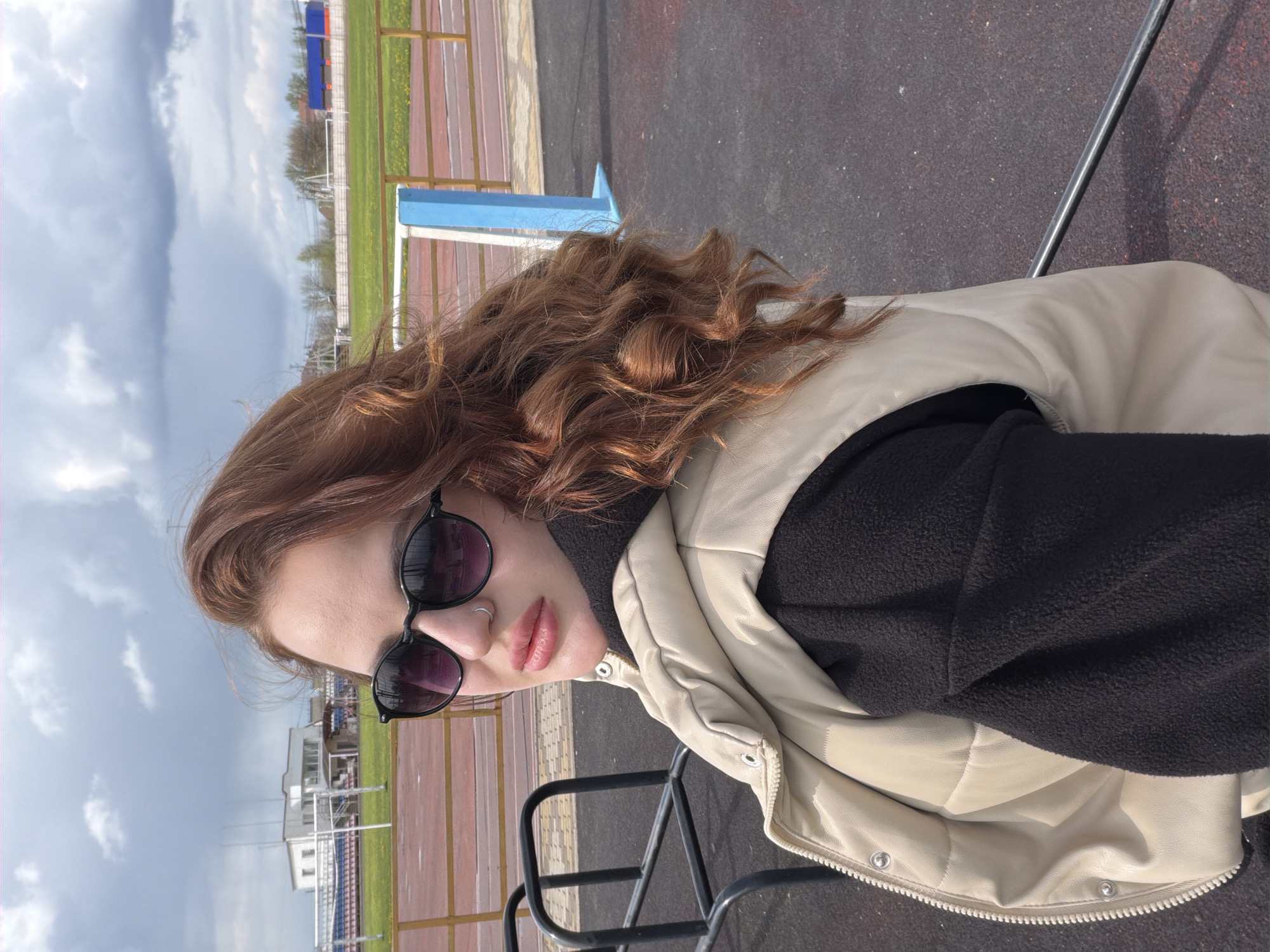 A woman with auburn hair and sunglasses is captured at an outdoor location, likely in Novopokrovskaya, Russia. The background reveals a sports field, characterized by a running track with red and brown lanes, a section of green grass, and blue and white metal railings. Additionally, a light-colored building is visible in the distance. The woman is dressed in a black garment underneath a puffy, light-beige jacket. The picture appears to be taken in daylight under a sky with patchy clouds.
