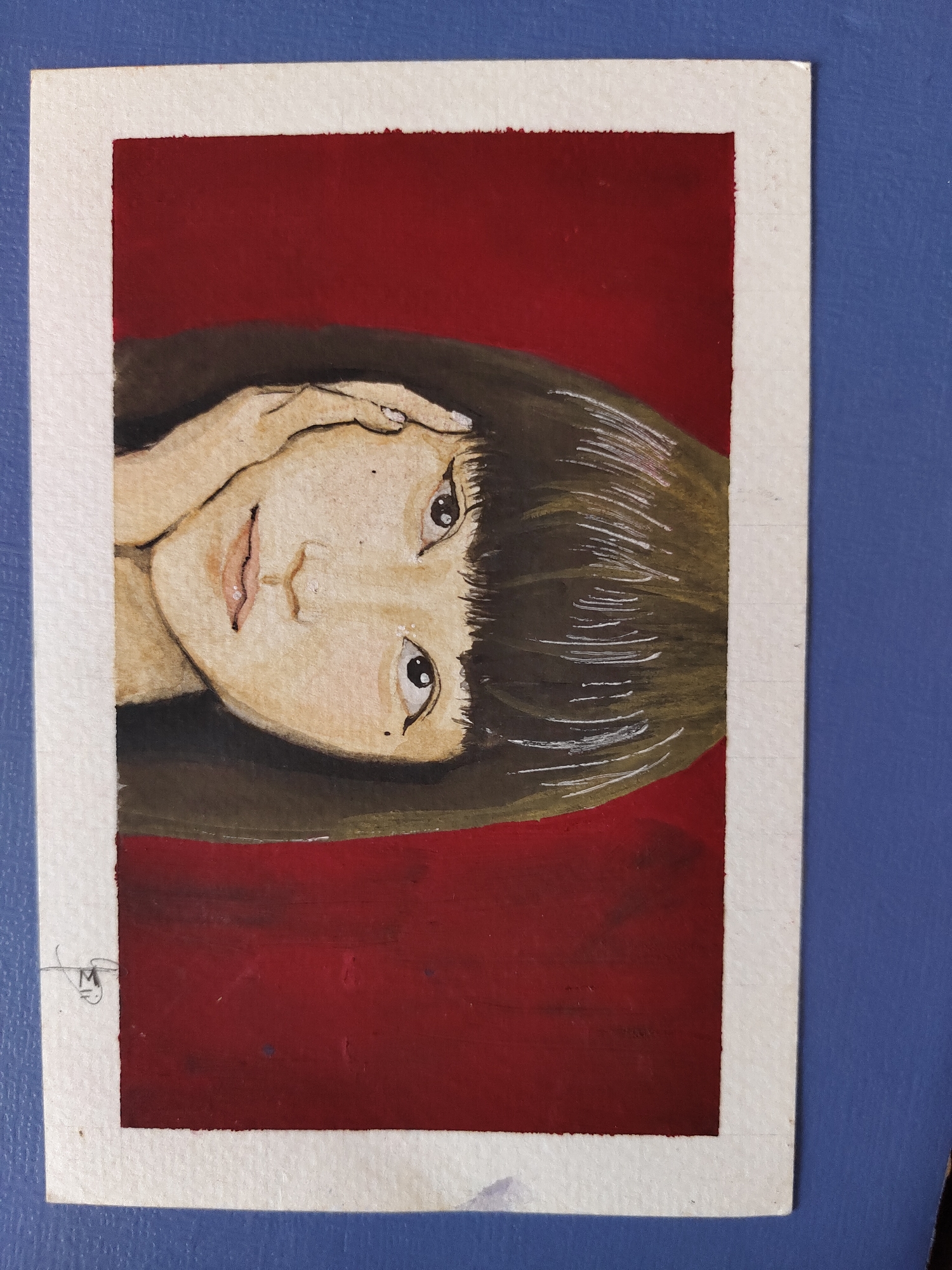 A stylized portrait painting, executed with what appears to be watercolor or gouache on paper, depicts a person's head and shoulders. The painting is oriented vertically but the subject's head is tilted 90 degrees to the viewer's left. The subject has fair skin, dark brown hair cut in a bob with bangs, and large, dark, expressive eyes looking slightly upwards. Their lips are parted, showing a hint of pink. A delicate hand with visible fingers rests gently against the forehead or hair, with a small white highlight suggesting a ring or light reflection on one finger. Small dark moles are visible below the left eye and near the right eye. The hair features fine white lines to indicate highlights.
The background of the portrait is a solid, rich, dark red, framed by a textured white border of the paper. The painting itself has visible brushstrokes, particularly in the hair and skin tones, adding to its artistic texture. A small, stylized signature, resembling an "M" with a horizontal line, is located in the bottom left corner of the white border.
The entire artwork is placed on a textured blue surface, possibly a fabric or board, creating a contrasting backdrop for the painting. The scene evokes a pensive or contemplative mood. The media file was captured in the context of Davao City, Philippines.