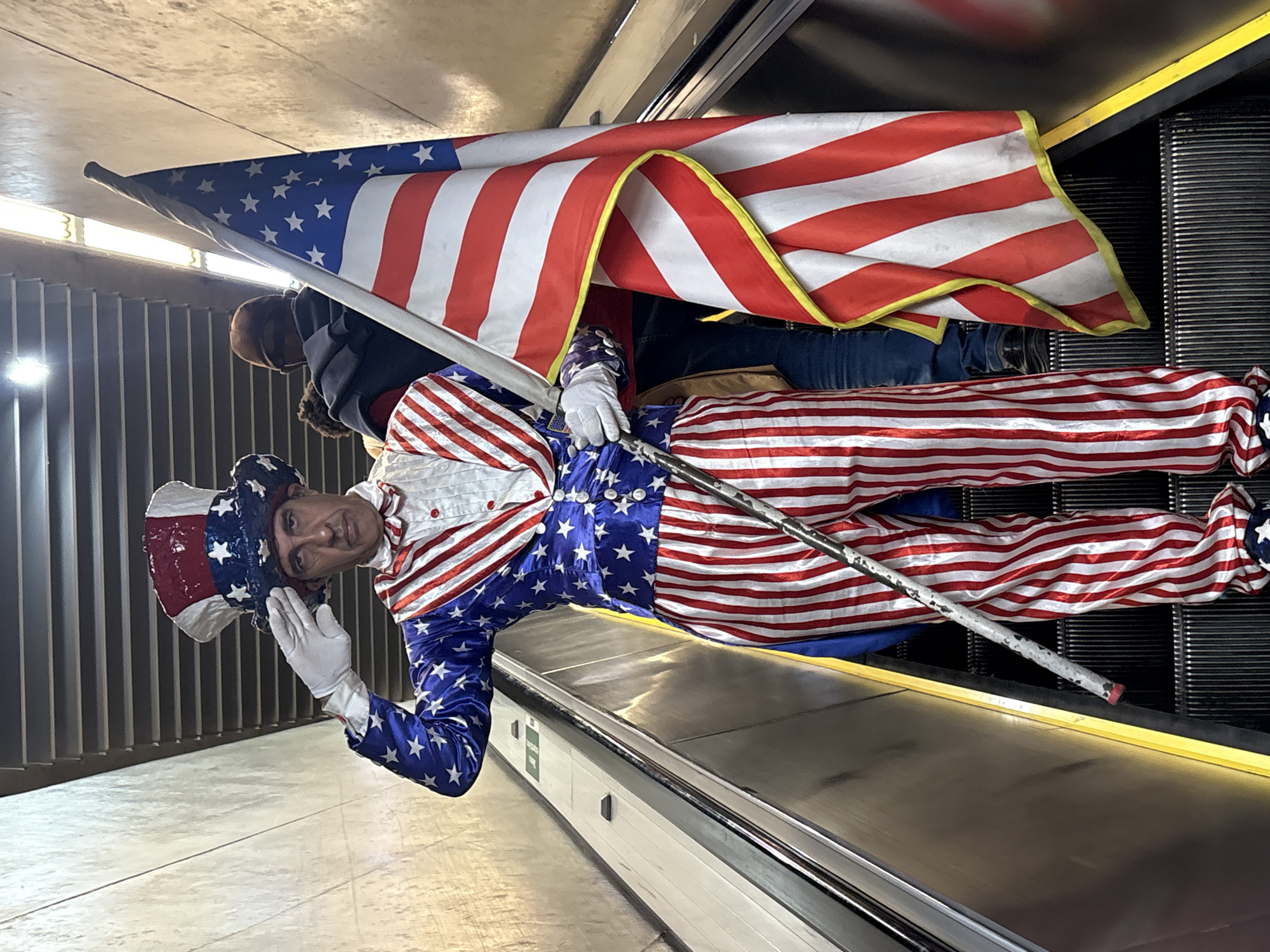 A man dressed as Uncle Sam stands on an escalator, holding an American flag and saluting. He wears a tall, red, white, and blue hat adorned with stars, a blue jacket with white stars, a white shirt with a red and white striped vest, and red and white striped pants. He also wears white gloves and holds a cane. Another person, wearing a brown hat and dark jacket, stands behind him on the escalator. The escalator appears to be indoors, possibly in a public transportation hub or shopping center, with metallic walls and fluorescent lighting. The scene suggests a public gathering or demonstration. The presence of the American flag and costume indicates a patriotic theme. The man's posture and gesture suggest a formal or respectful acknowledgment. The specific location in São Paulo, Brazil is not identifiable from the provided image.