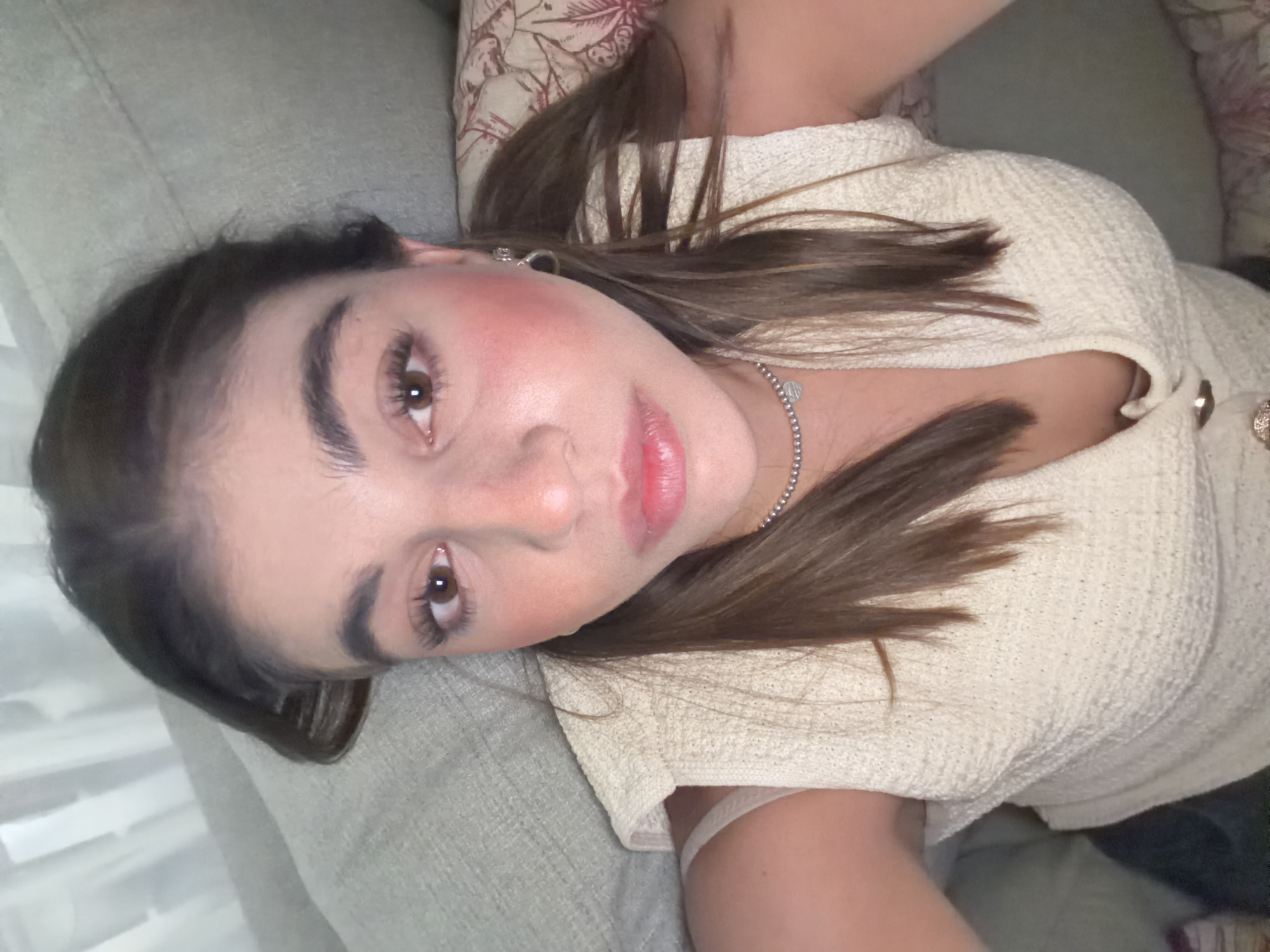 A young woman is lying on a gray couch, her head resting on a pillow with a floral pattern. She is wearing a light-colored, textured top and a silver necklace. She has brown hair and brown eyes. Her complexion is fair, and she appears to be wearing makeup, including blush and lip color. The location is likely indoors, and the image is taken from a slightly elevated angle, looking down on her. The background is softly lit, revealing a hint of sheer curtains. There is no information to place the image at any specific address in Barrio de Nuevo León, Mexico, or any other location.