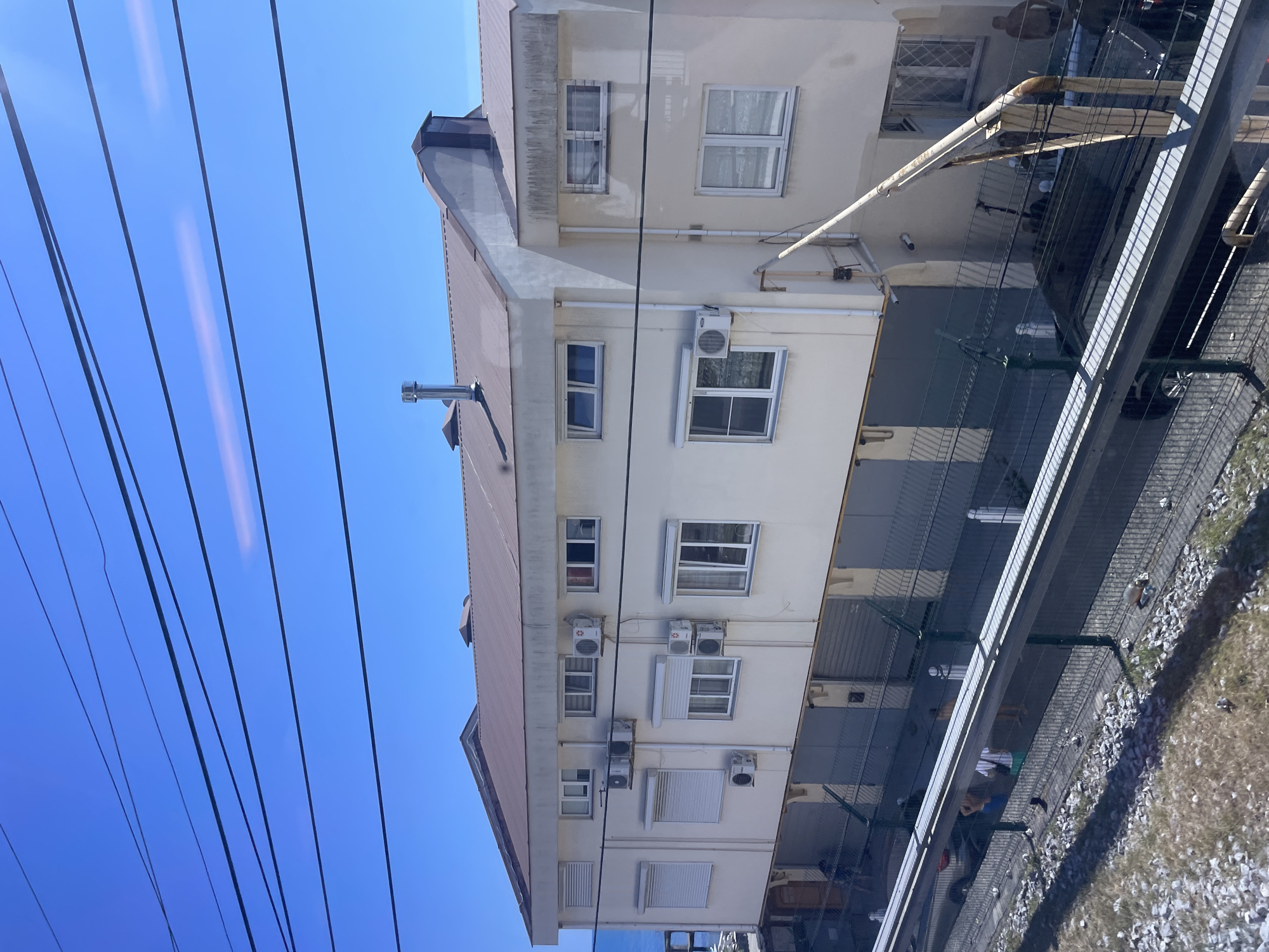 The image shows a multi-story building with a brown roof and cream-colored walls. The building has numerous windows, some with blinds. Several air conditioning units are mounted on the exterior walls. There is a metal chimney on the roof, as well as other ventilation pipes.
In the foreground, there is a chain-link fence with a metal barrier above it. Beyond the fence, there are several vehicles, including a dark-colored car, and a few people are visible, some appearing to be standing near the garages. The ground appears to be a mix of gravel and dry grass.
In the background, the blue sea is visible, suggesting a coastal location. The sky is clear and blue. The overall scene depicts a utilitarian building, possibly part of a commercial or industrial area, near the sea. The presence of overhead wires across the frame indicates urban infrastructure.
While the image depicts a building and its surroundings, there is no specific visual information within the image to definitively identify the location as Sochi, Russia. The image is a general depiction of a coastal urban or semi-urban environment.