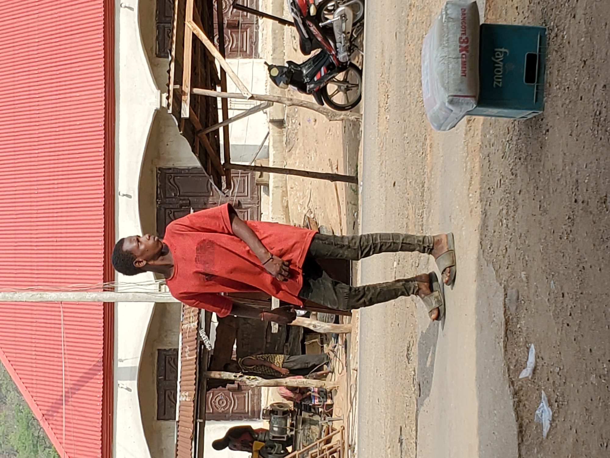 This outdoor photograph captures a street scene, likely in Mararaba, Nigeria, under bright daylight. The image is oriented vertically but depicts a horizontal ground level.
In the foreground, a young man stands upright, facing to his right. He wears a red short-sleeved t-shirt, dark trousers or jeans, and open-toed sandals. His expression is neutral as he looks into the distance. His hands are clasped loosely in front of him.
Behind him, on the left, is a building with a prominent red corrugated metal roof. The building itself is light-colored, possibly white or cream, with dark brown wooden doors and archways visible beneath a wooden support structure. Further back, near the center, a motorcycle with red and black accents is parked on an unpaved, dusty ground. Another individual, possibly a child, is partially visible sitting on a raised wooden platform under a makeshift shelter. Near the bottom of the frame, another person is seen by some machinery, which could be an engine or pump.
On the right side of the frame, near the foreground, there's a light-colored, possibly paved, surface transitioning into a dusty, unpaved area. A white bag of "DANGOTE 3X CEMENT" rests on top of a green plastic crate, which has the brand name "Fayrouz" visible on its side. Some discarded white plastic litter is scattered on the ground further right. The overall setting suggests a bustling, semi-urban environment.