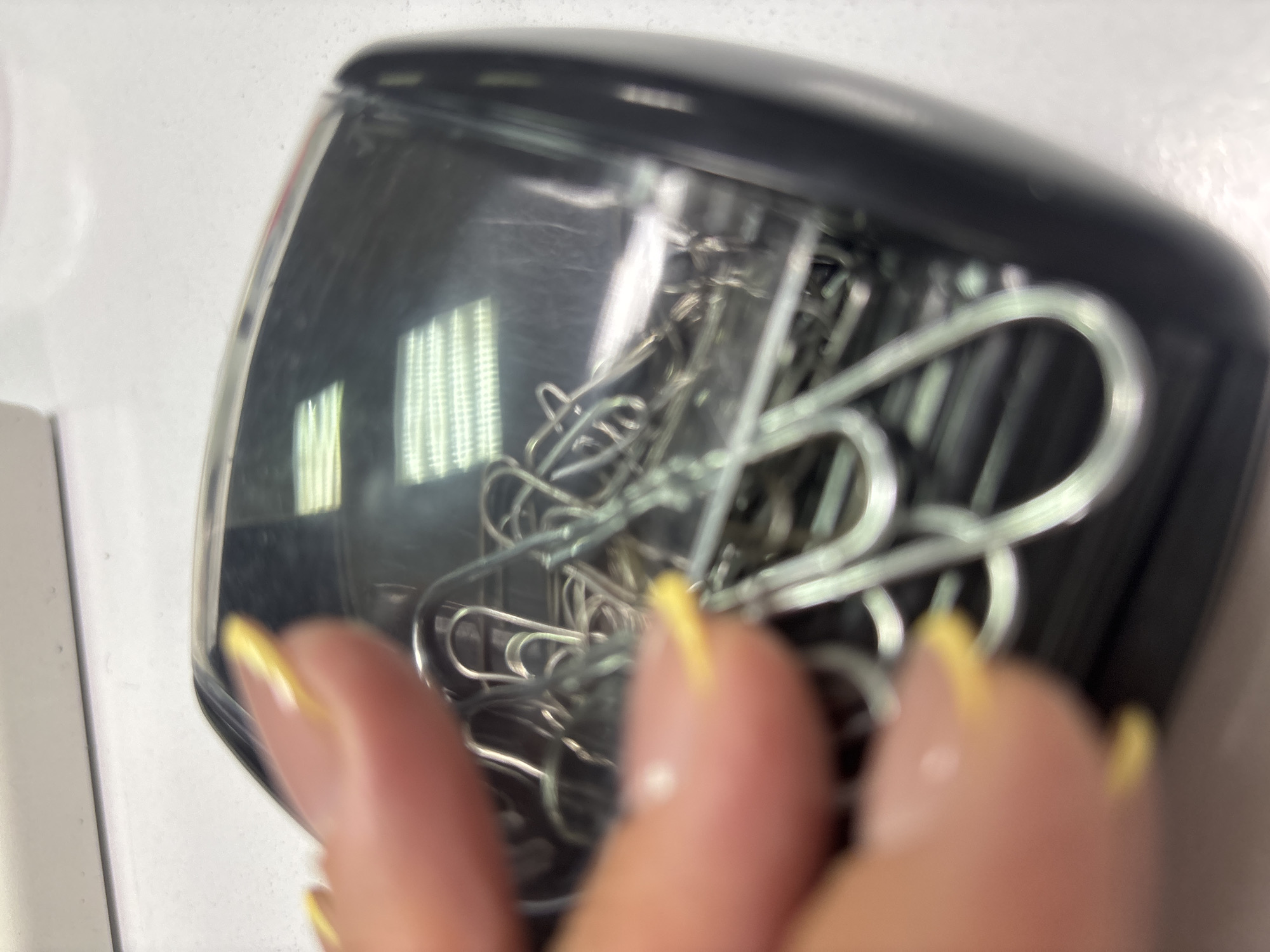 A close-up photograph shows a black, clear-lidded container filled with an assortment of silver-colored paper clips. A human hand, partially visible in the foreground, is positioned near the container. The fingernails are painted with a light yellow color. The background is a plain, white surface. The specific location (city of Chaikovsky, Russia) is not identifiable from visual information alone.