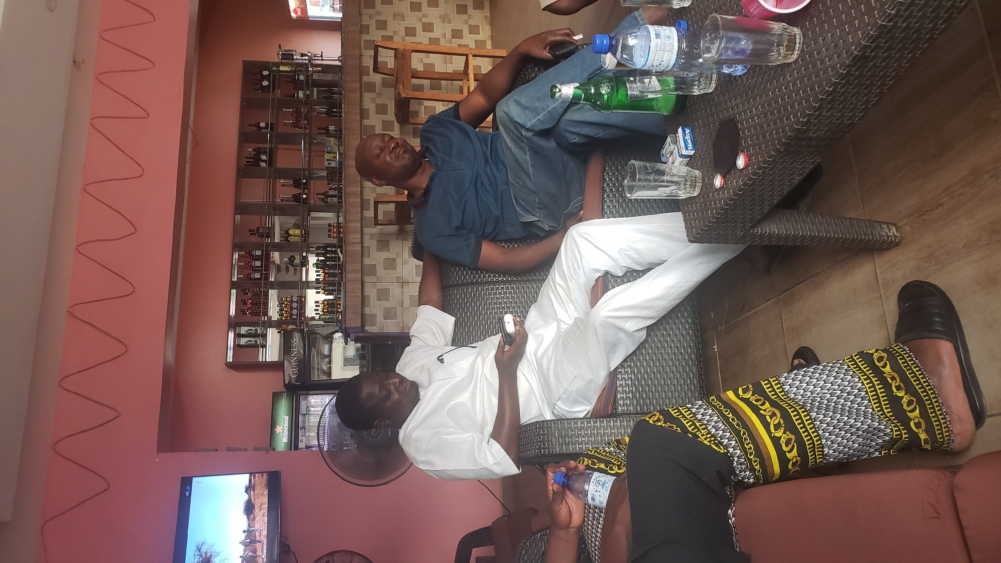 The image shows three men relaxing in what appears to be a bar or lounge setting. One man is reclined on a sofa, dressed in a blue shirt and jeans. Another is reclining on the same sofa, wearing a white shirt and pants. A third man is seated beside the sofa, clad in black pants and yellow patterned fabric on his legs.
A low table with drinks and glasses sits in front of them. The background includes a wall with liquor bottles displayed on shelves. A television screen is visible on another wall.
