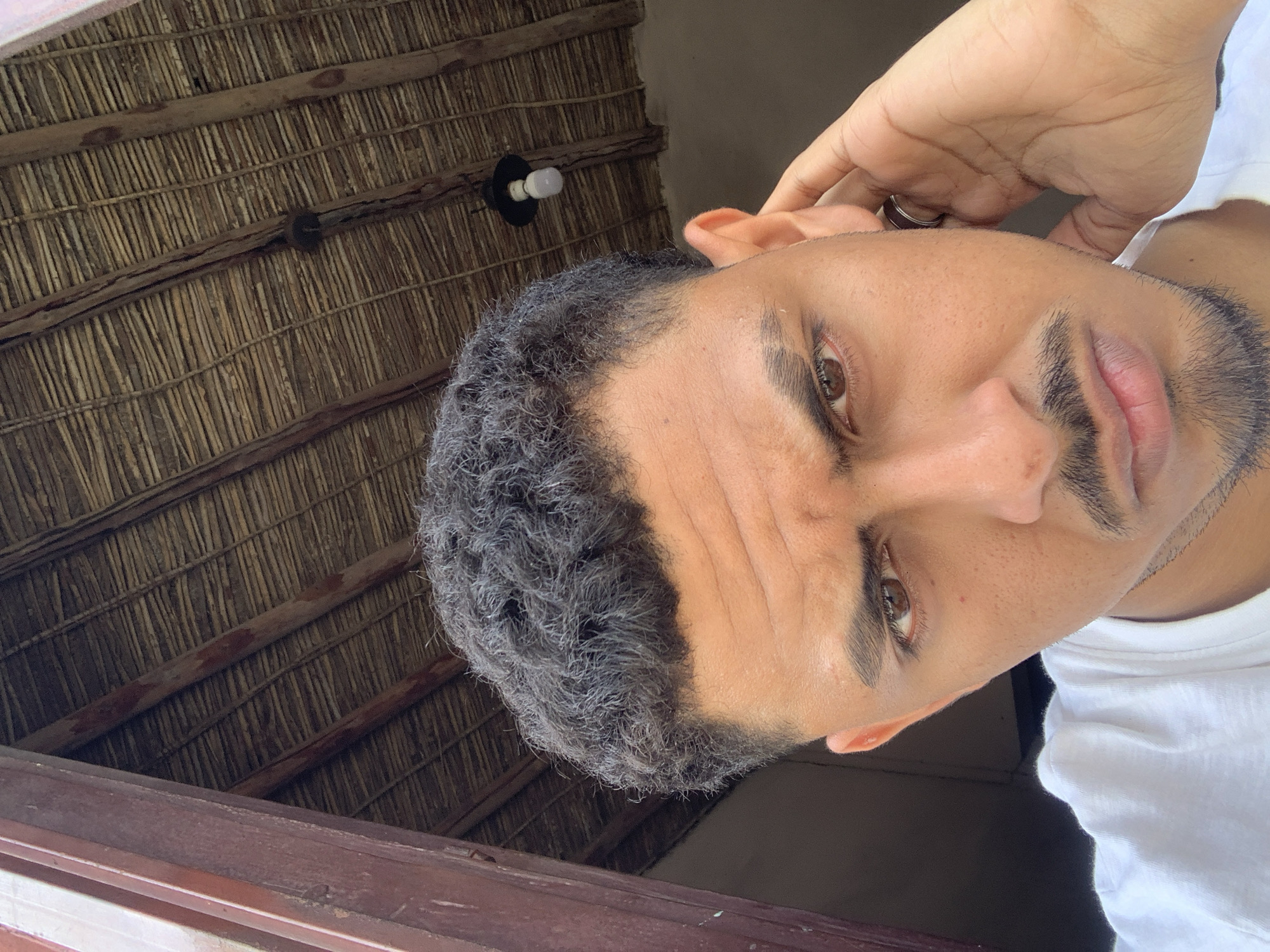 A young man with curly dark hair, a mustache, and a goatee poses for a selfie indoors, likely in Santa Ana, Venezuela, where the rustic thatched roof ceiling suggests a rural or traditional setting. He wears a white t-shirt and has his right hand raised to his ear. The lighting is natural and comes from the right.