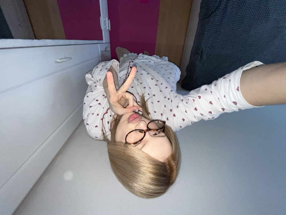 A fair-skinned person with blonde hair and glasses poses, wearing a white pajama top with red hearts. They display a peace sign near their face. The photo is taken at an odd angle, with the person seemingly lying down. Visible in the background are a white cabinet, a pink wall, wooden surfaces, and part of a blue surface, potentially a couch or a blanket. The floor is pale, and the overall lighting suggests an indoor setting.