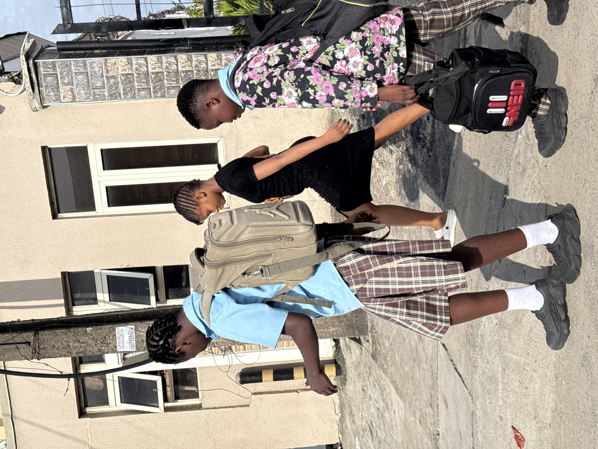 Three young people walk along a sidewalk in Ado, Nigeria, alongside a light-colored building. The person in the foreground wears a light blue shirt, plaid skirt, black sneakers with white socks, and carries a tan backpack. The person in the middle wears a black dress and white sneakers. The person in the background wears a floral patterned shirt, plaid pants, black sneakers, and carries a black backpack with red and white lettering. The building has several windows, and a utility pole is visible in the background.