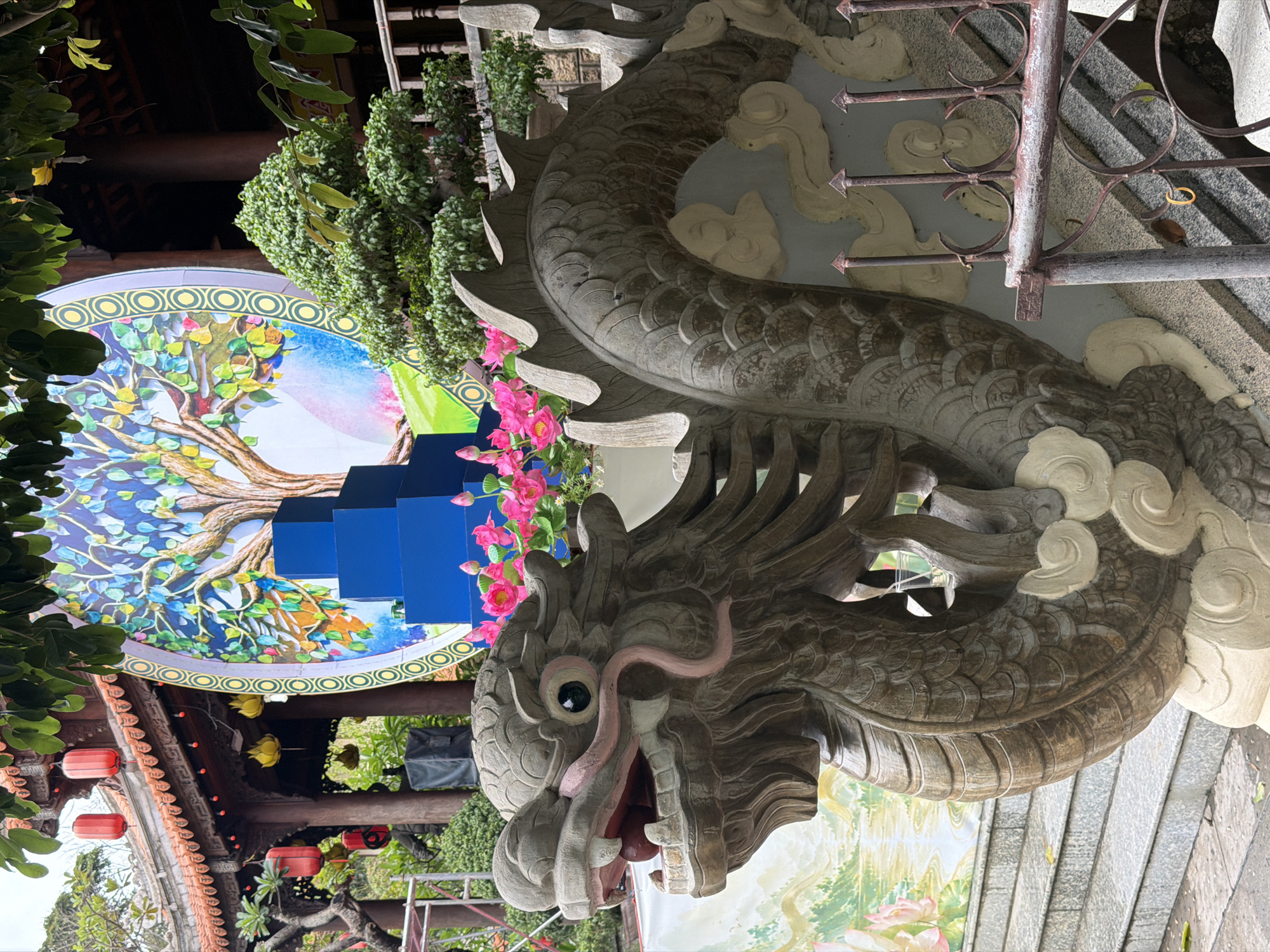 A stone dragon sculpture is positioned near stairs in Tay Nha Trang Ward, Vietnam. The dragon's head is facing the viewer. Behind the dragon are decorations including a tree art piece, blue steps, and pink flowers. Red lanterns are visible on the roof of a building to the left. A grey metal railing appears above the dragon's back.