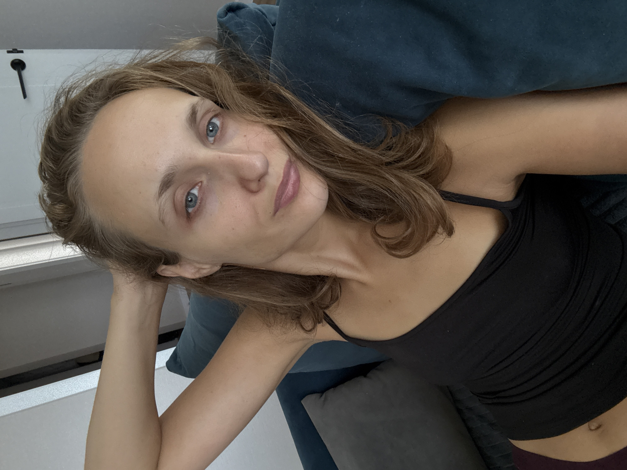 A Caucasian woman with light brown hair, blue eyes, and fair skin is captured in a selfie. She is lying on a teal sofa, with her left arm bent behind her head. Her gaze is directed at the camera. The background shows a grey pillow on the sofa and a door with a black handle on the right. The lighting suggests an indoor environment. The exact location in the city of Подшивалово, Russia, cannot be determined from the visual information provided in the image.