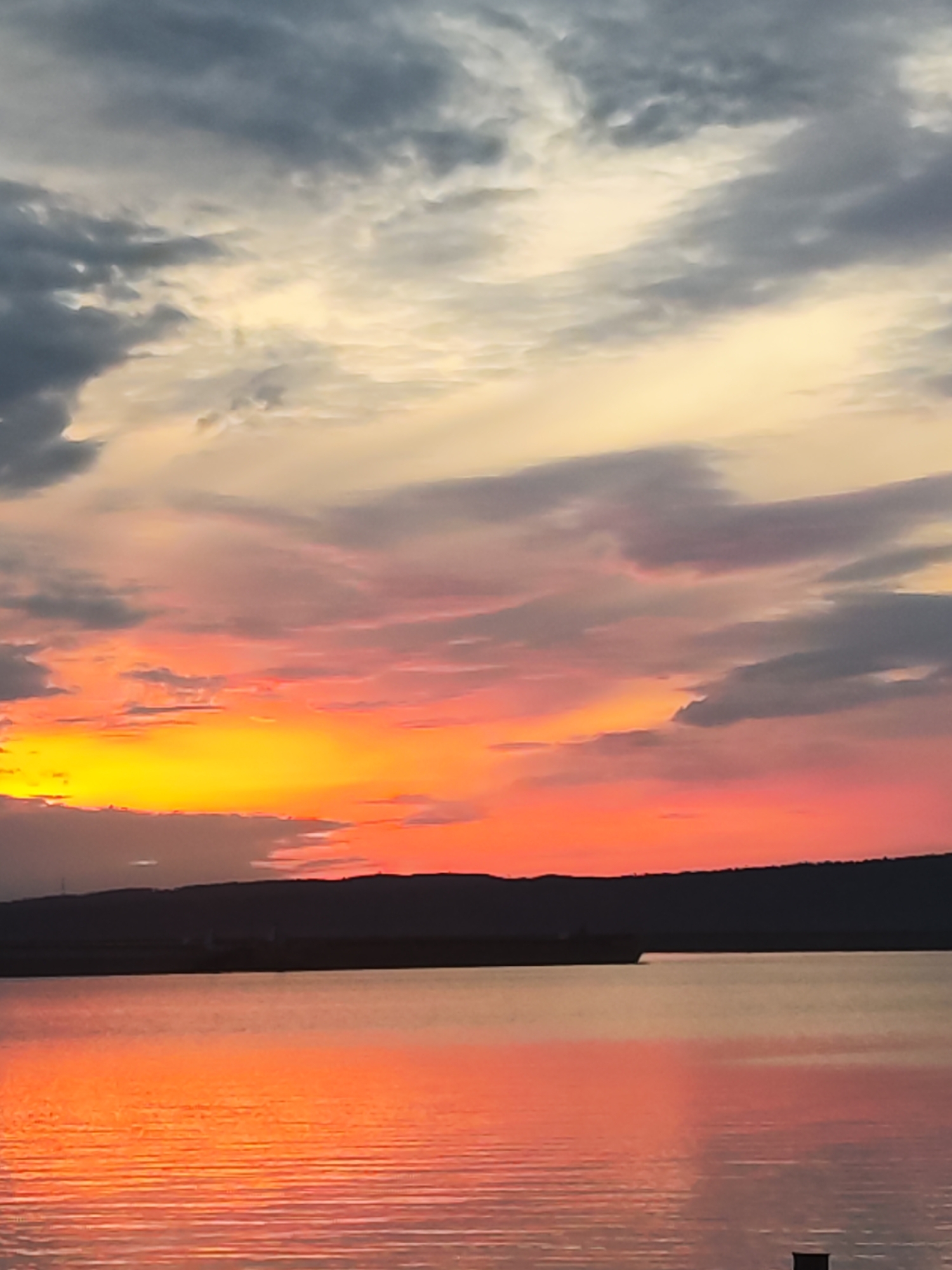 The image depicts a sunset over a body of water, with land visible in the middle ground. The sky is filled with clouds, with colors ranging from yellow and orange near the horizon to pink and gray higher up. The water reflects the colors of the sunset, creating an orange glow. The land appears as a dark silhouette against the bright sky. The location is likely near Chaikovsky, Russia, based on the geographical characteristics of the landscape, including a wide river and some rolling hills. The scene is calm and serene.
