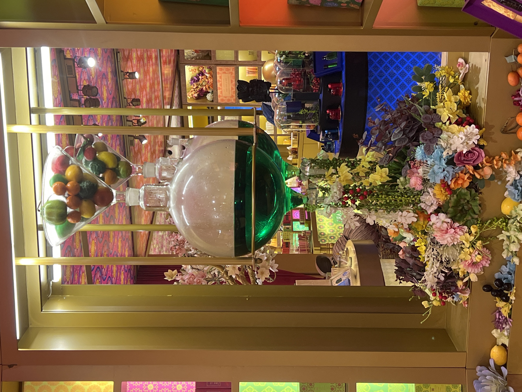 The image depicts a brightly lit display window, likely within a retail establishment in Galle, Sri Lanka. The window features a tiered arrangement of decorative objects. At the top, a glass cone-shaped vessel contains a colorful assortment of artificial fruits, including green, orange, and red spherical items. Below this, two large, clear glass spheres, possibly part of a distillation apparatus, hold a vibrant green liquid. The spheres are suspended and adorned with artificial flowers and greenery, creating an opulent, botanical effect.
The background of the display is a riot of color and pattern. The ceiling is adorned with a vibrant pink and gold patterned fabric. Behind the central display, a mirrored surface reflects the elaborate ceiling pattern and what appears to be a store interior with shelves displaying various products. The shelves are filled with small bottles and boxes, suggesting a perfumery or cosmetic shop.
In the foreground, a collection of artificial flowers in various shades of pink, yellow, blue, and red are arranged in a lush bouquet. Smaller decorative elements, such as artificial fruits (oranges and grapes) and purple flowers, are scattered at the base of the display. The overall impression is one of a luxurious and visually rich product display, aiming to attract customers with its elaborate presentation. The colors are saturated, and the lighting is intense, highlighting the textures and details of the displayed items.