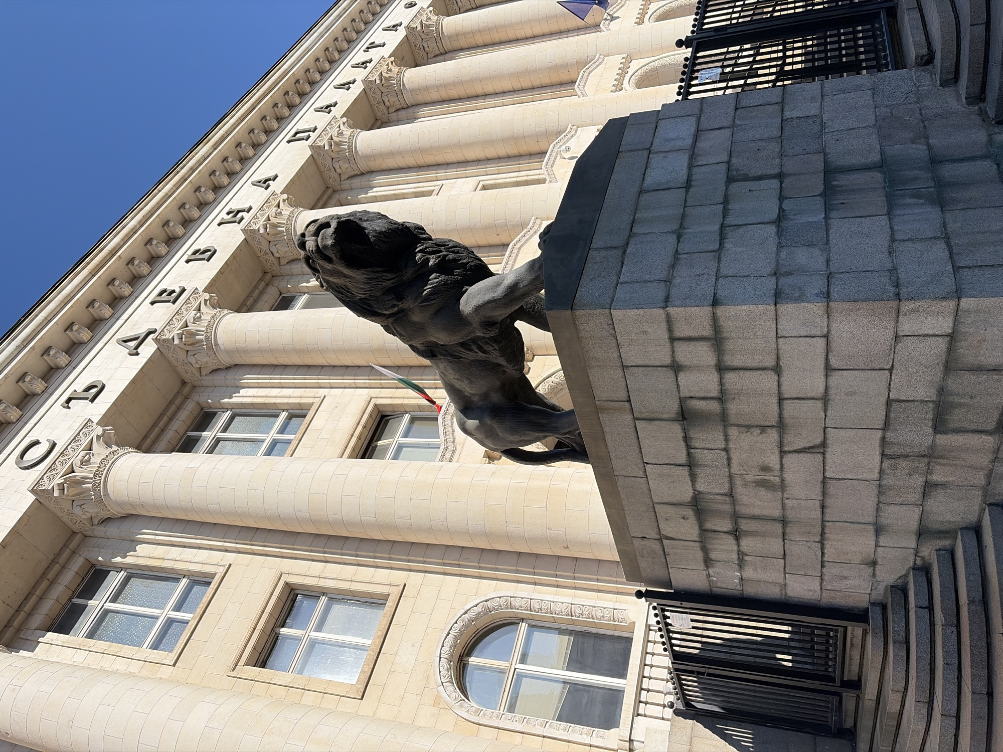 The image shows a statue of a lion in front of a large, ornate building in Sofia, Bulgaria. The building, which is likely a government or official institution, features classical architecture with large columns, arched windows, and decorative stonework. The lion statue is dark in color and appears to be made of bronze or stone. It stands on a raised stone pedestal. A flag, appearing to be the Bulgarian flag with its white, green, and red stripes, is visible behind the lion. In the background, the sky is clear and blue. The perspective is low-angle, looking up at the lion and the building. The scene is set on a bright, sunny day.
