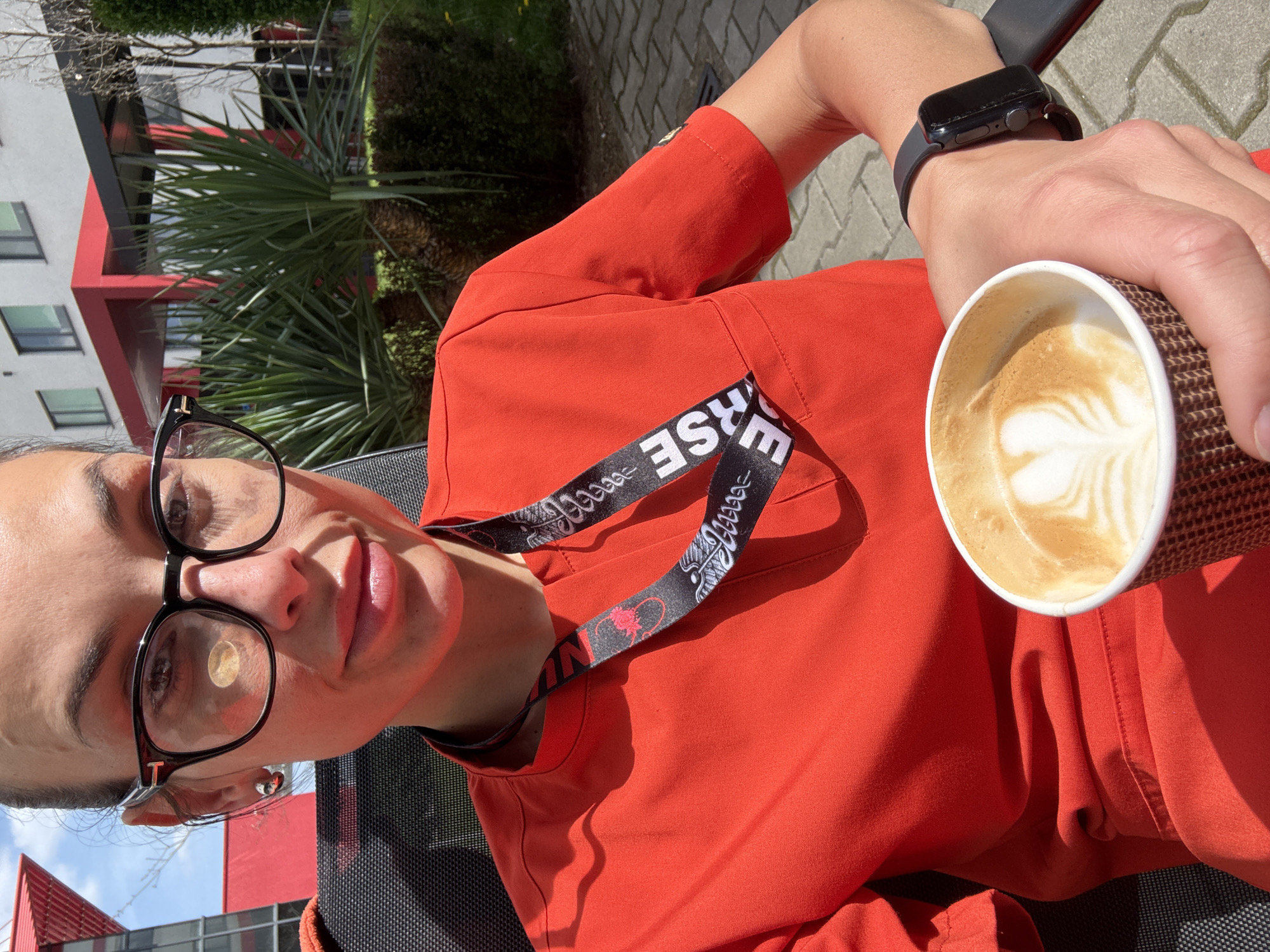 A person is depicted relaxing outdoors in Pleven, Bulgaria, enjoying a cup of coffee on a sunny day. The individual, appearing to be a woman, wears black-framed glasses and an orange-red top. A black lanyard, partially showing the text "VERSE" and decorative patterns, hangs around her neck. On her left wrist, she wears a dark smartwatch. She holds a disposable coffee cup in her right hand, which contains a beverage with a detailed latte art pattern on its foam.
The scene is set in an urban environment with strong sunlight creating clear shadows, suggesting it's either morning or early afternoon. In the background, modern buildings with red and light-colored panels are visible, along with green foliage, possibly palms or yucca plants, and the branches of a deciduous tree. The ground is paved, and the person is seated in a dark mesh chair. The overall impression is one of calm and leisure.